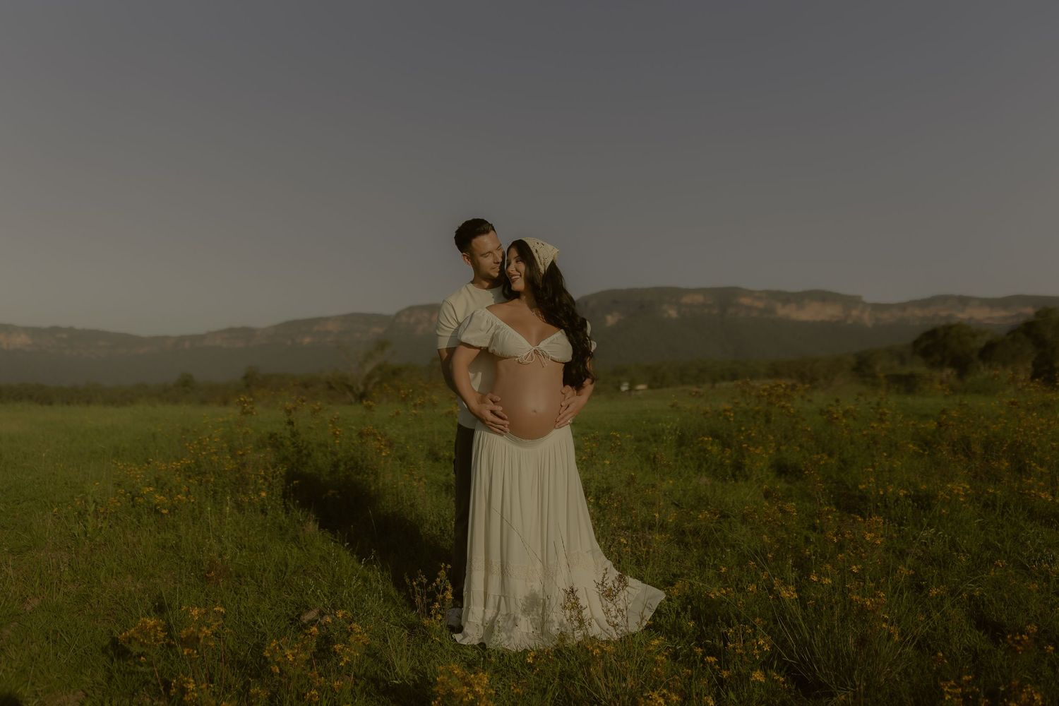 A couple embraces in a white dress against a scenic mountain backdrop during golden hour in a wildflower meadow.