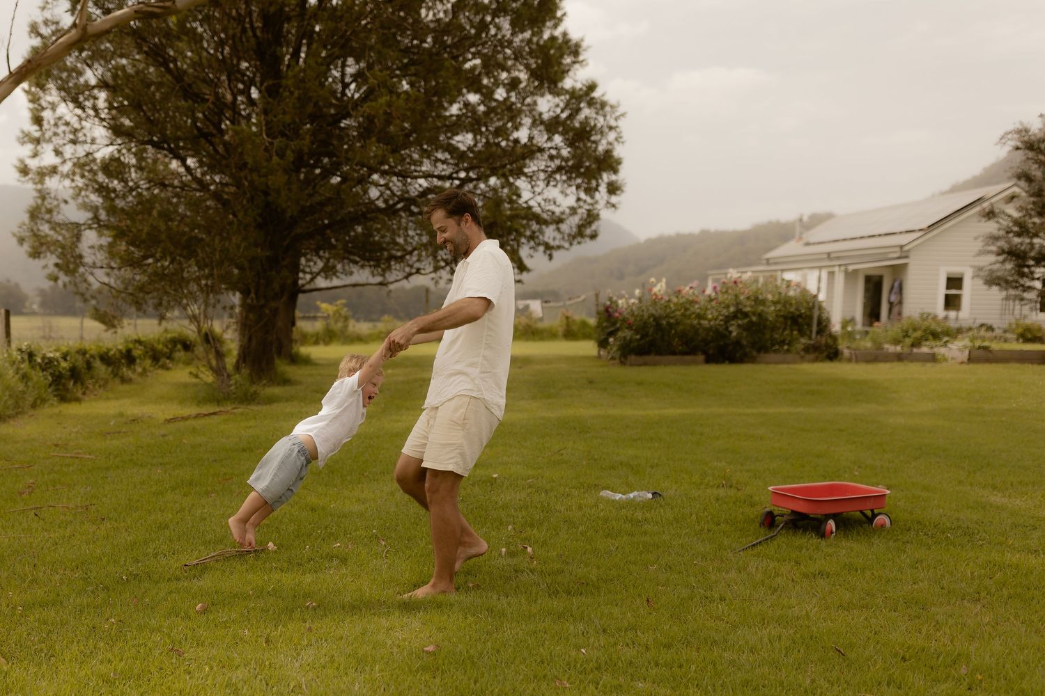 People playing and running in a grassy field with a red wagon and rural farmhouse in the background.