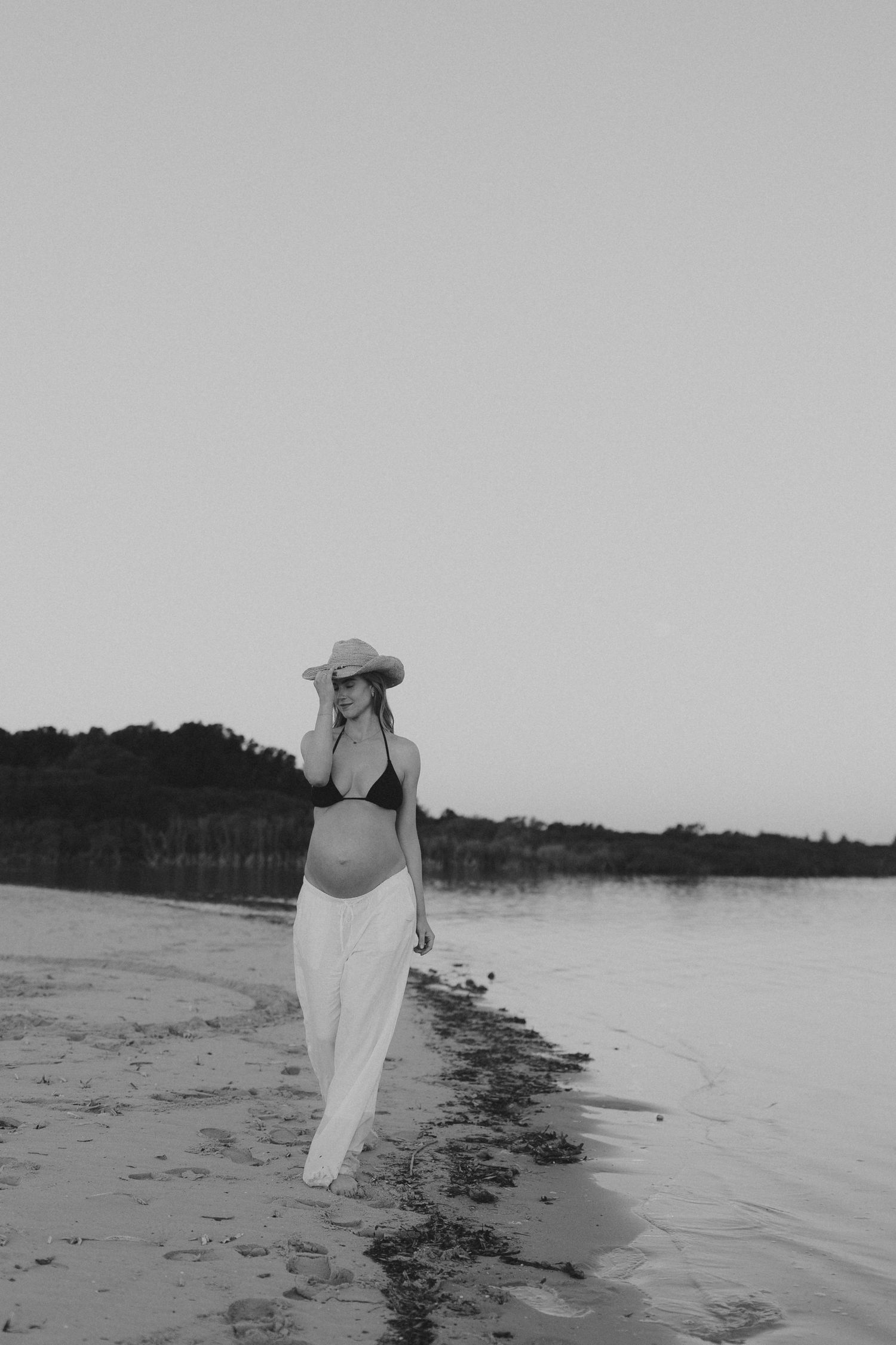 Black and white image of pregnant woman in bikini top, white skirt and sun hat walking along calm beach shoreline.
