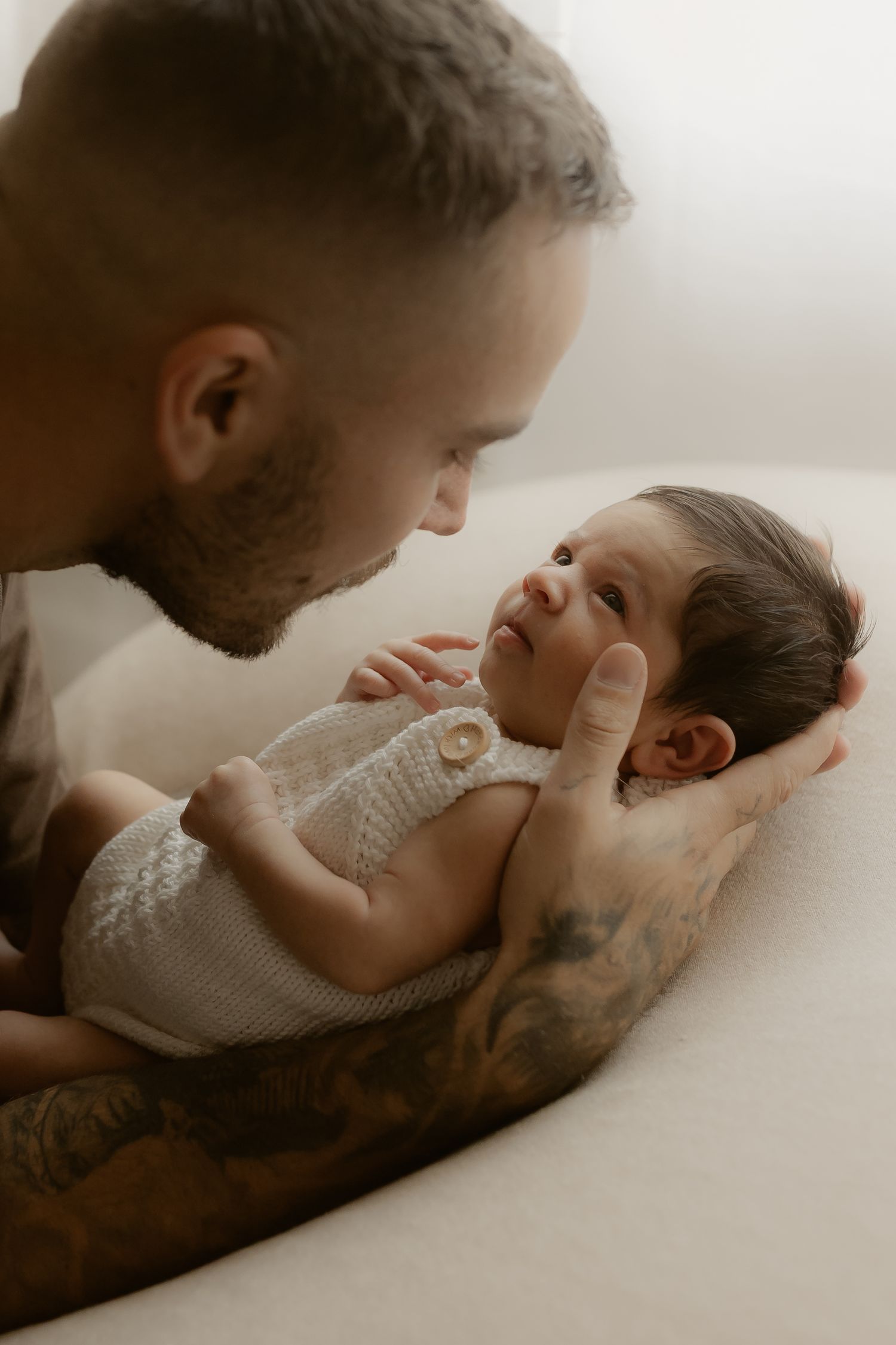Tattooed father gazing lovingly at his newborn baby dressed in a white knit romper, cradled in his arms.