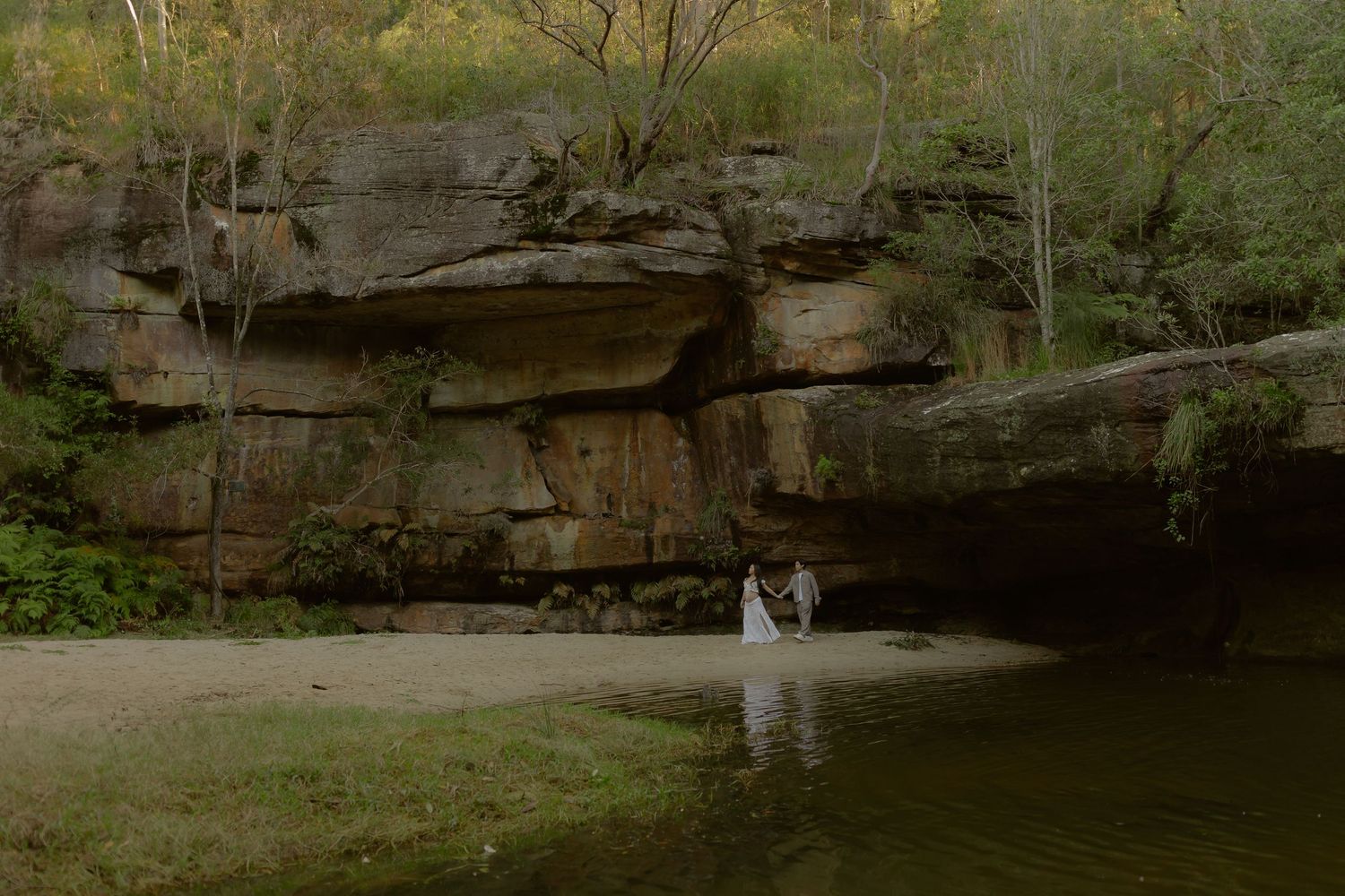 Couple in wedding attire standing by a serene river beneath dramatic sandstone cliffs surrounded by lush greenery.