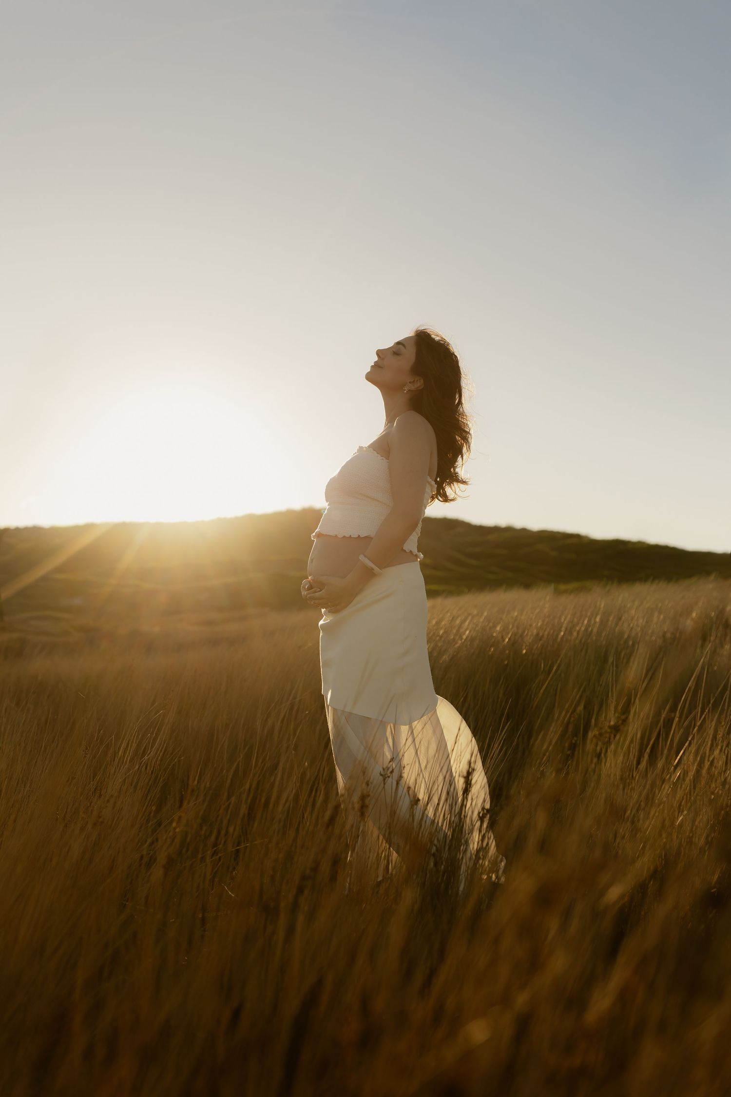 Pregnant woman in white off-shoulder dress standing in golden grass field at sunset.