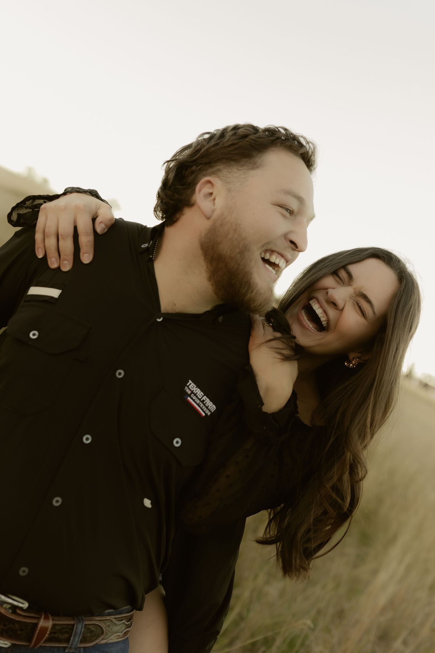 A joyful couple laughs together outdoors in a golden field, man wearing a black western shirt.