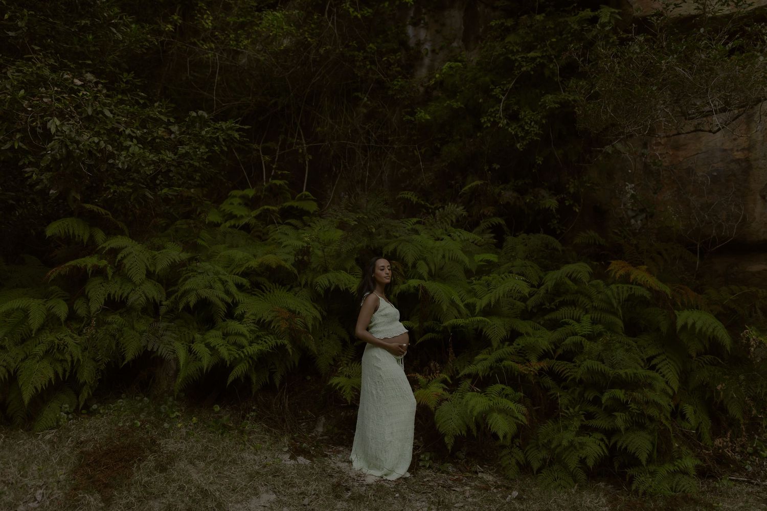 Pregnant woman in white dress standing among lush green ferns in a dark forest setting.