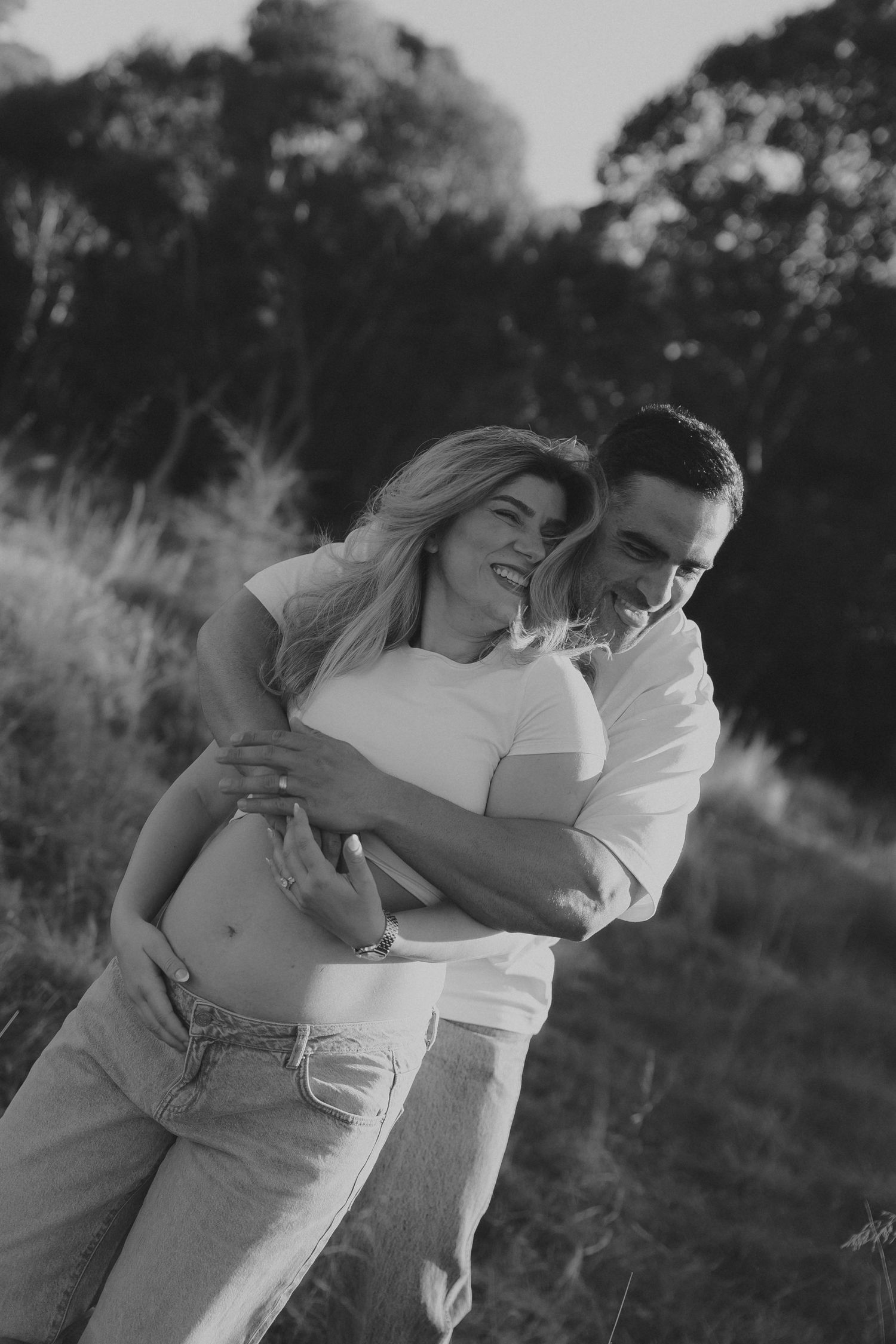 Black and white photo of a couple embracing outdoors, man hugging pregnant woman from behind, both smiling.