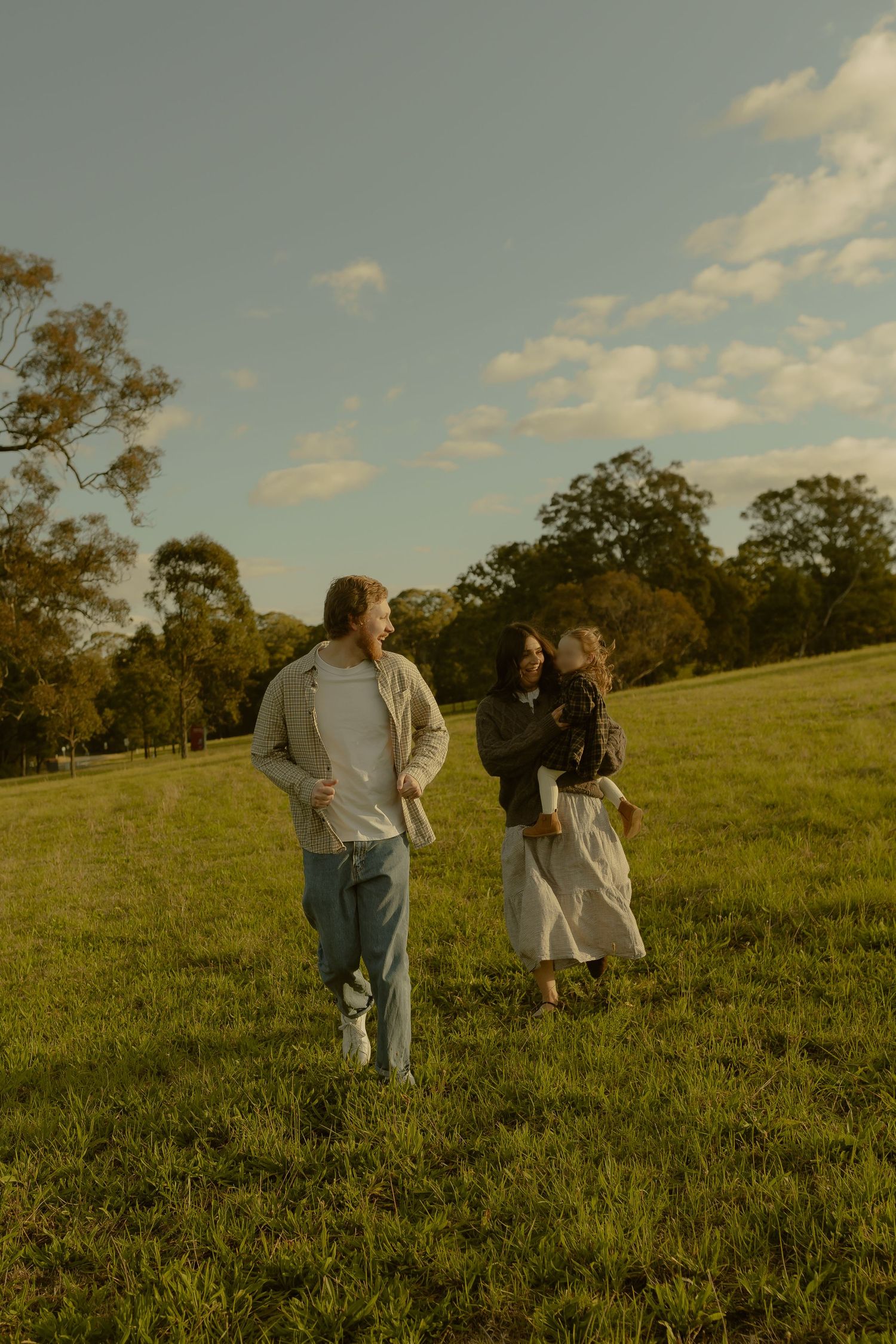 A couple walking and laughing together in a sunny green field with trees and blue sky in the background.
