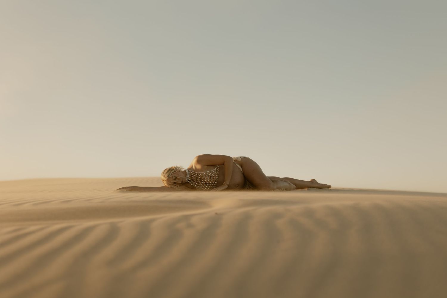 A person lying curled up on golden sand dunes in a vast desert landscape under a hazy sky.
