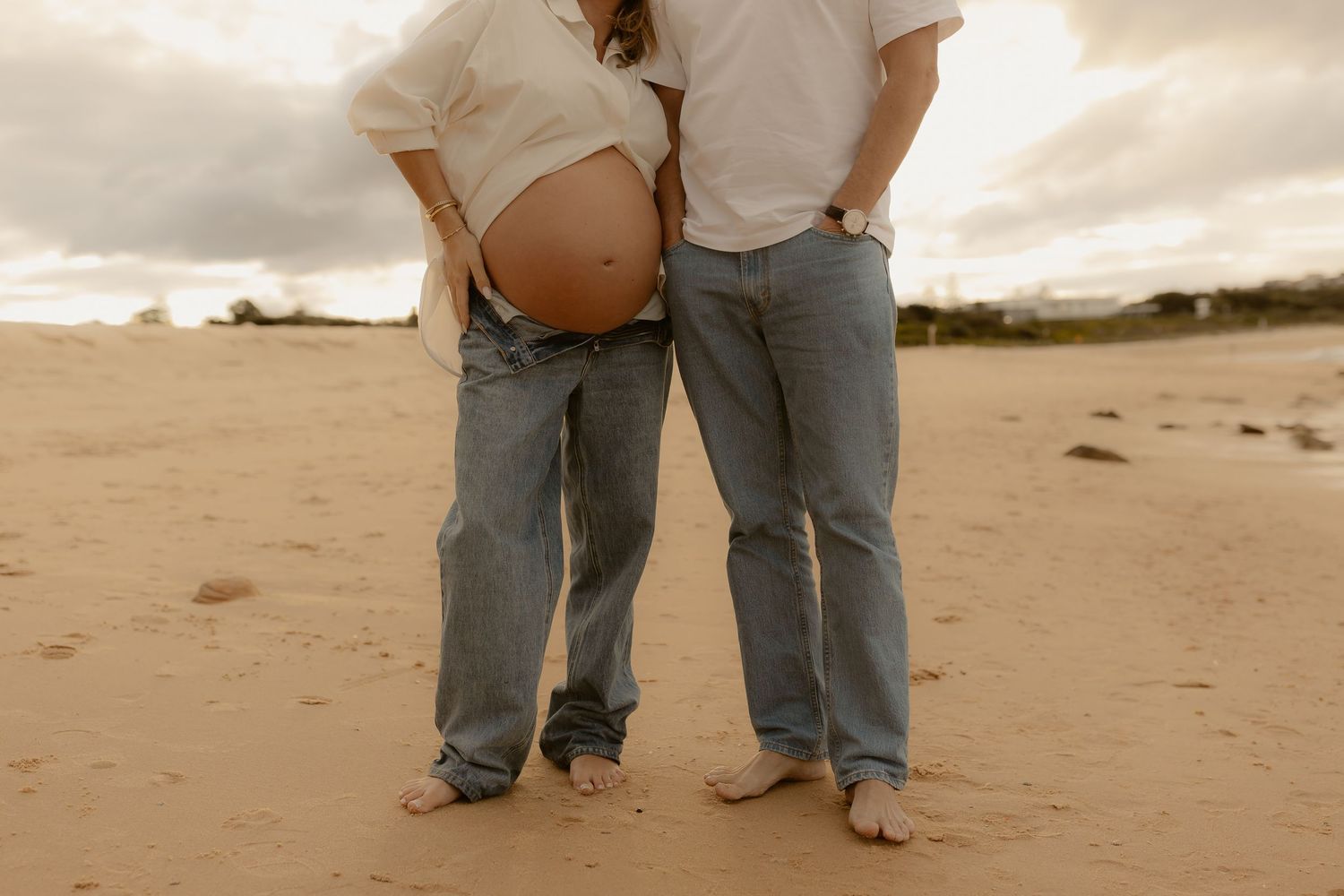Pregnant couple standing barefoot on sandy beach, woman showing baby bump, both wearing jeans and white tops.
