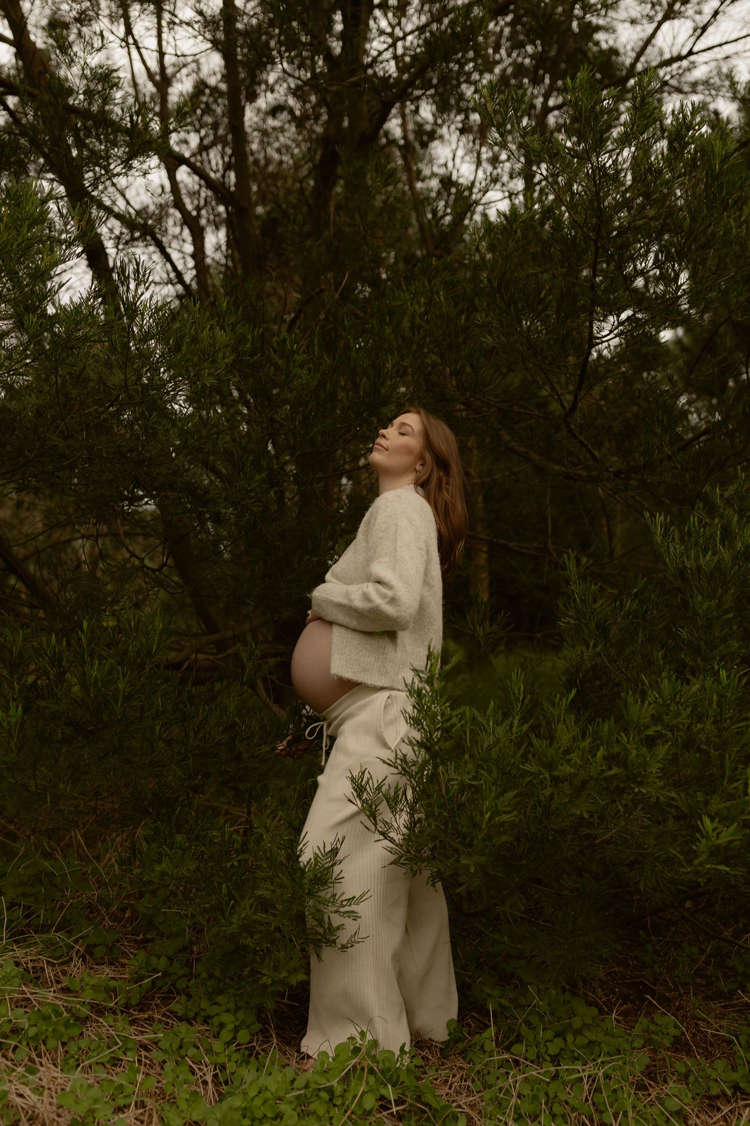 Pregnant woman in cream outfit cradling bump among green bushes, head tilted back.