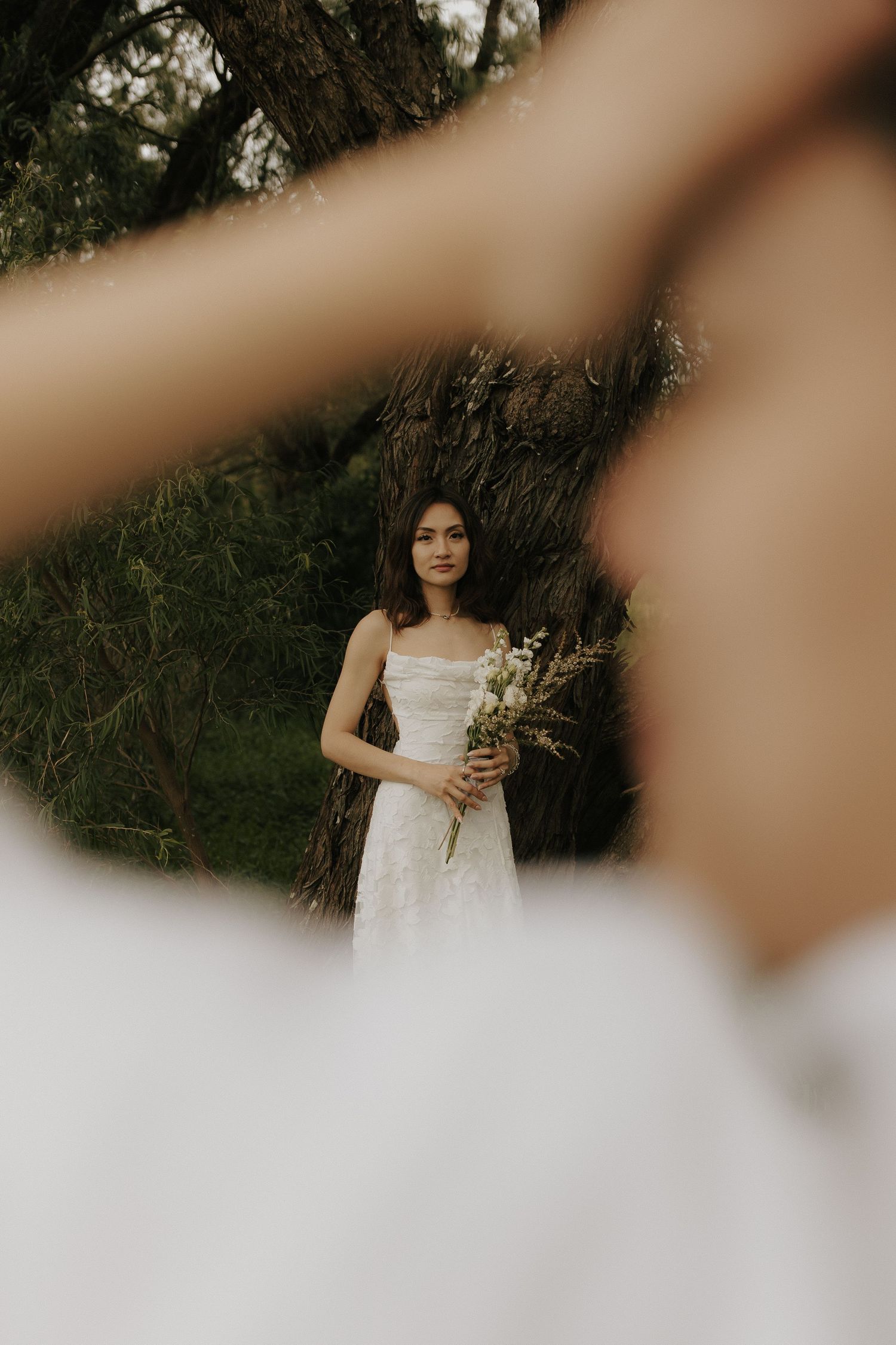 Artistic portrait through blurred foreground elements showing person in white dress holding flowers.