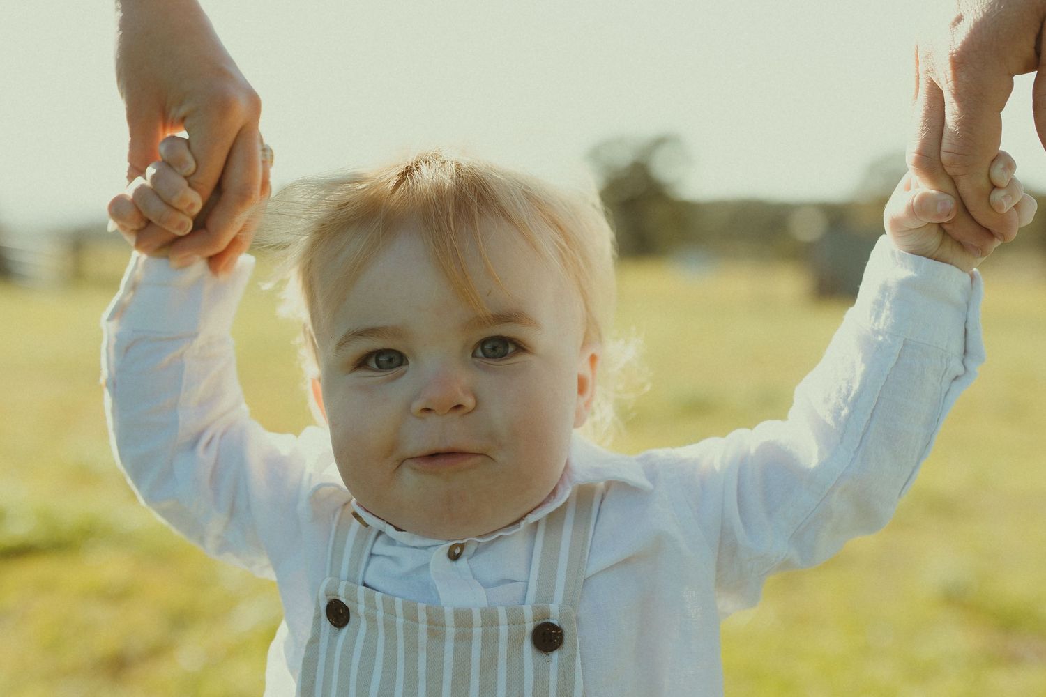 A small child in a white sweater raises their arms joyfully while playing outdoors in golden sunlight.