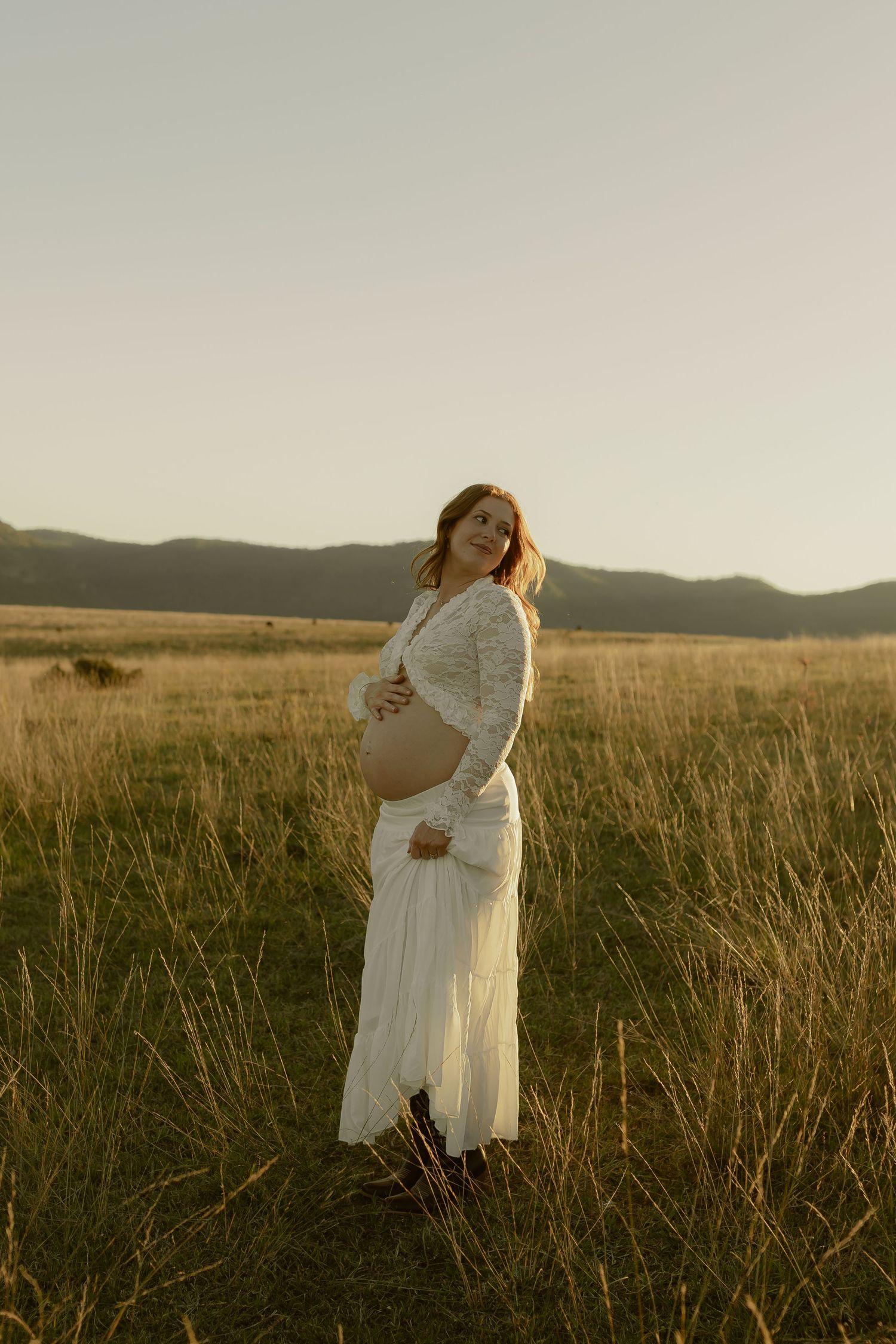 Pregnant woman in white dress holding baby bump in golden field at sunset with mountains in background.