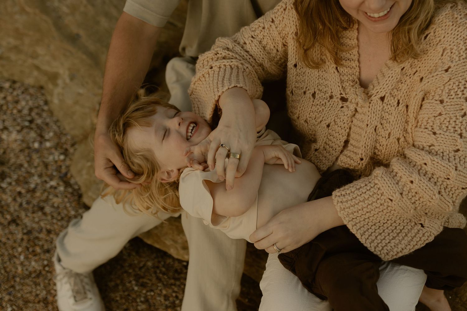 A joyful toddler laughs while being tickled by parents in neutral tones, captured in a warm family lifestyle photo.
