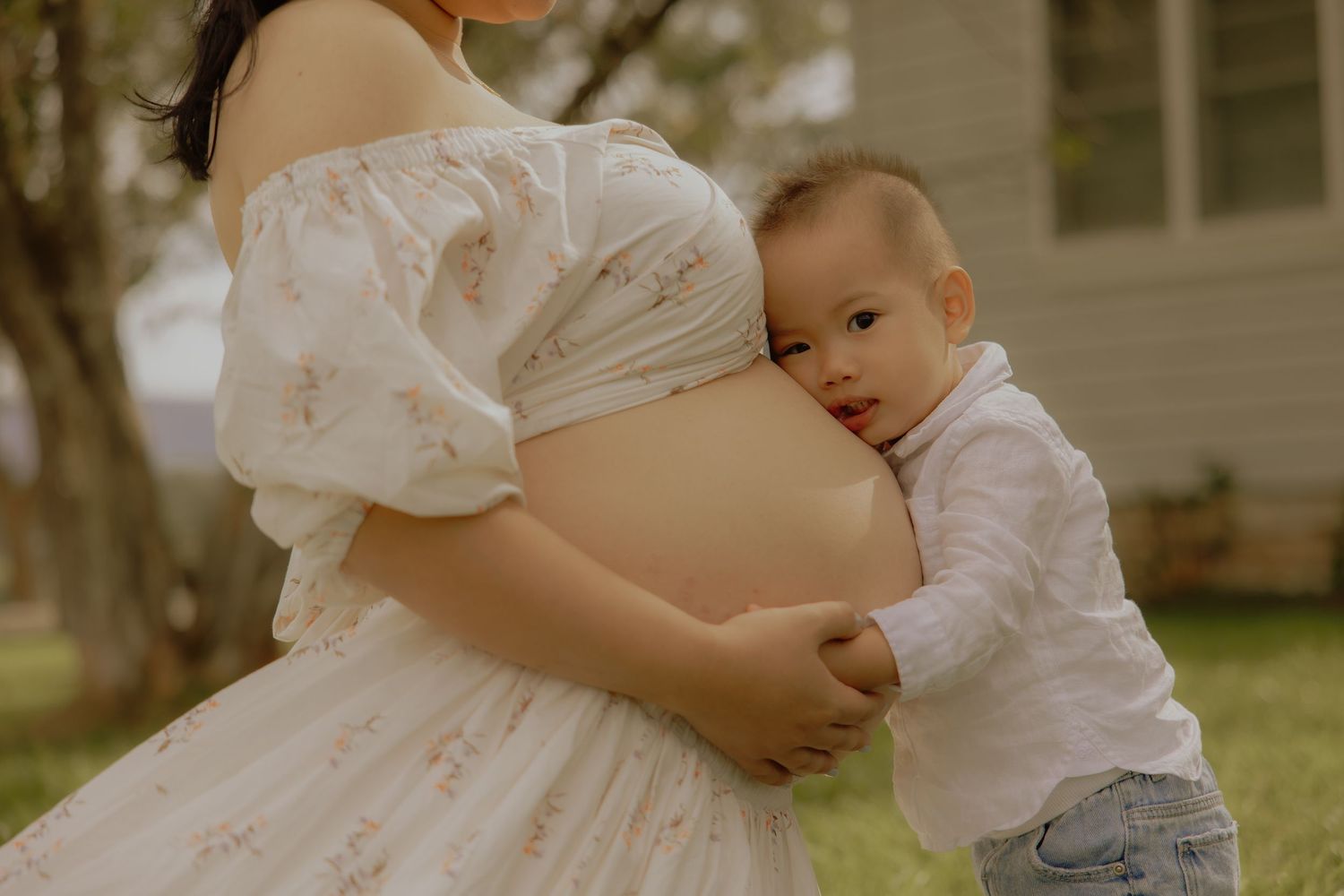 A toddler hugs a pregnant woman's belly outdoors, wearing white outfits in a heartwarming maternity photo.