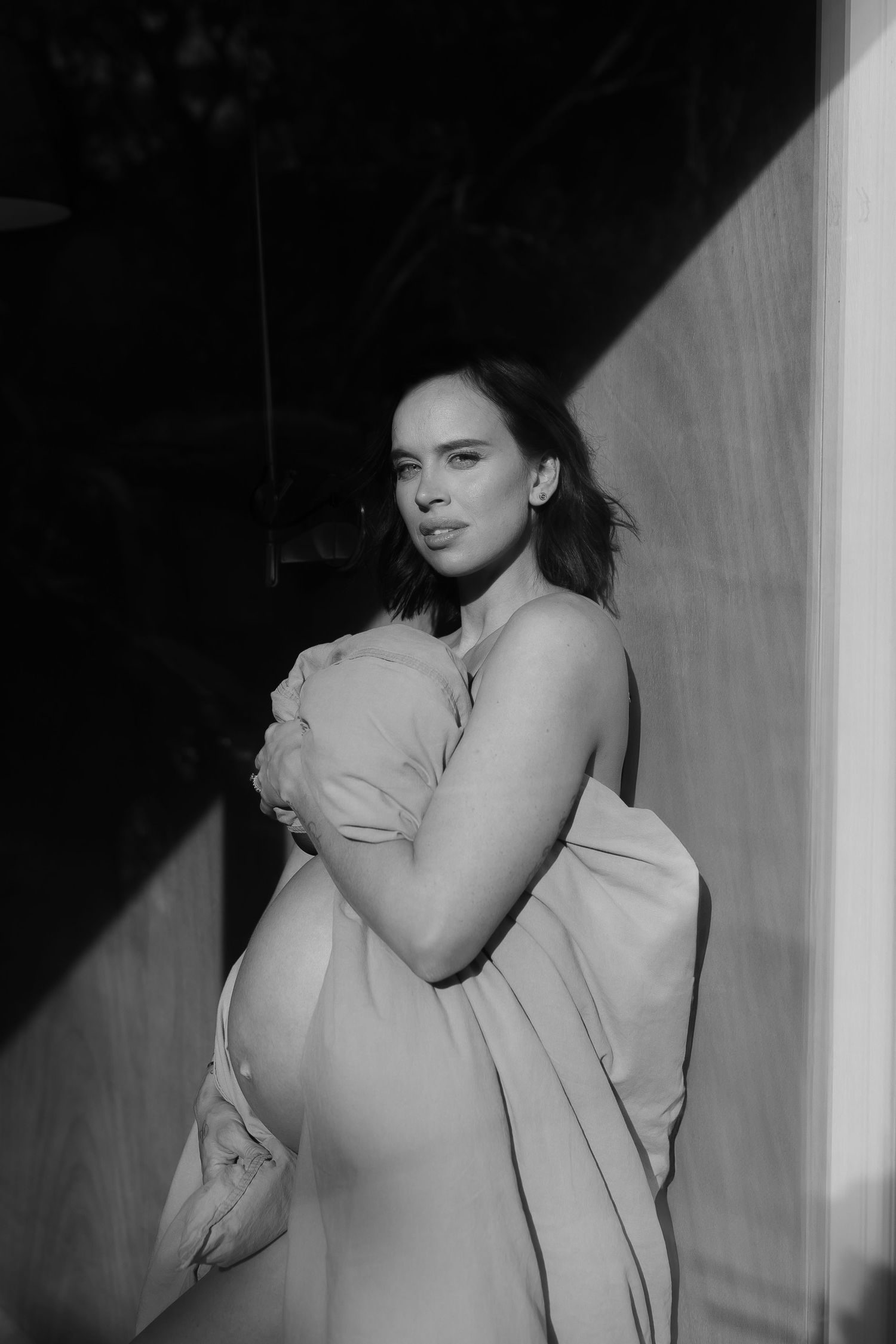 Black and white artistic portrait of an expectant mother draped in light fabric standing against a stark wall.