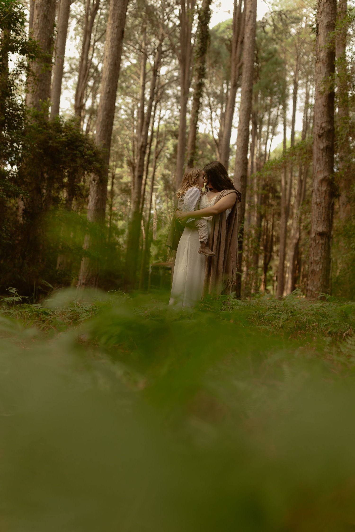 A couple embraces romantically in a lush green forest, surrounded by tall trees and ferns.