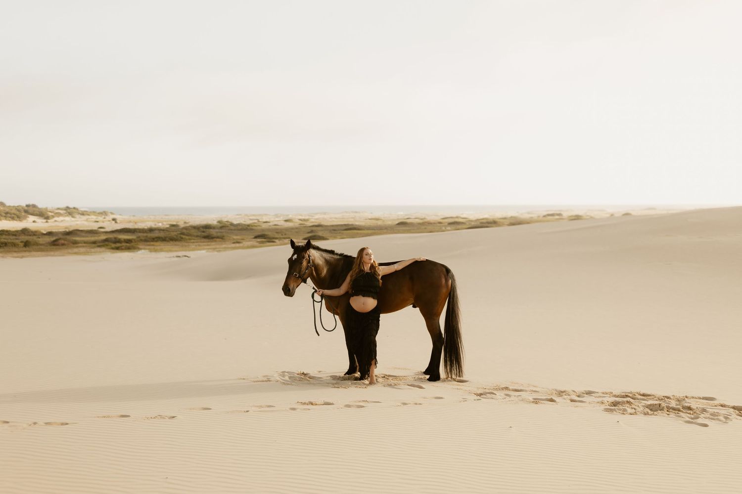 Silhouettes of a horse and person stand together on vast sand dunes during golden hour.