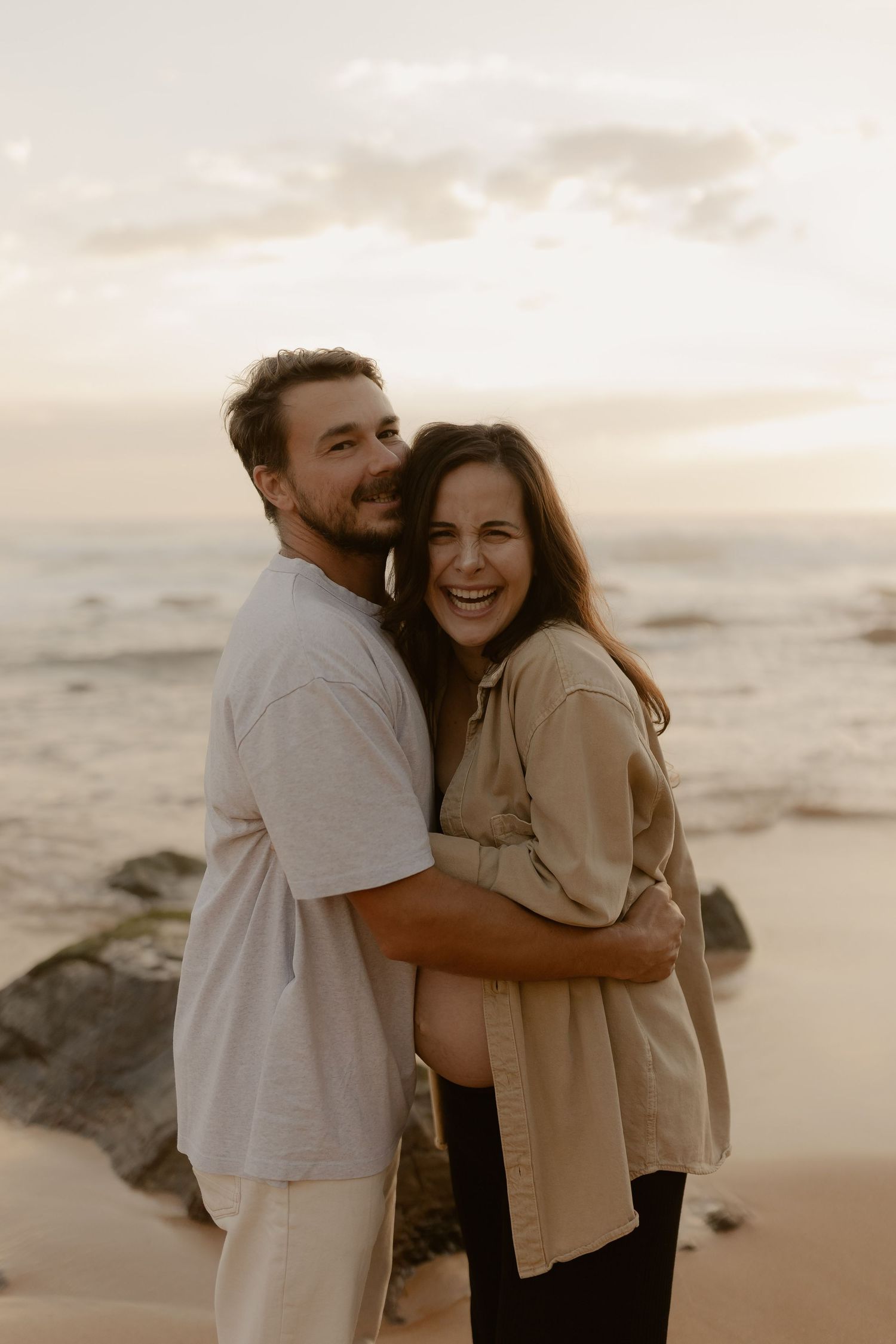 Two people embrace and laugh together on a beach at golden hour with ocean waves in the distance.