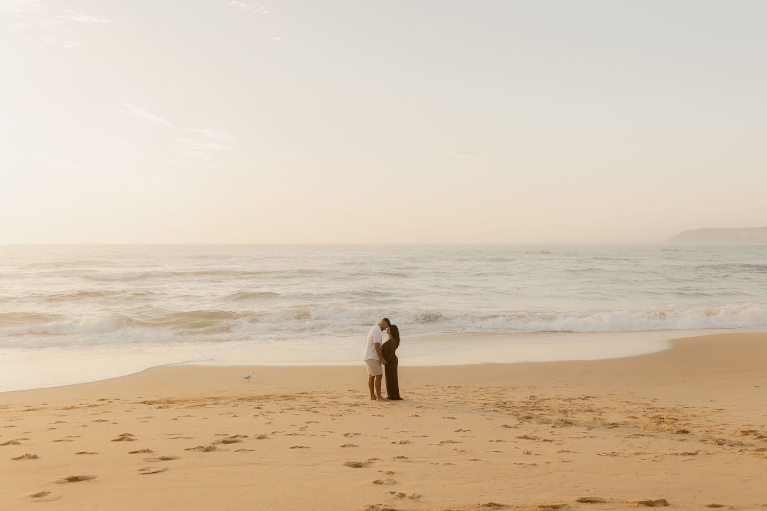 Two figures embracing on an empty beach during golden hour with misty ocean waves behind them.