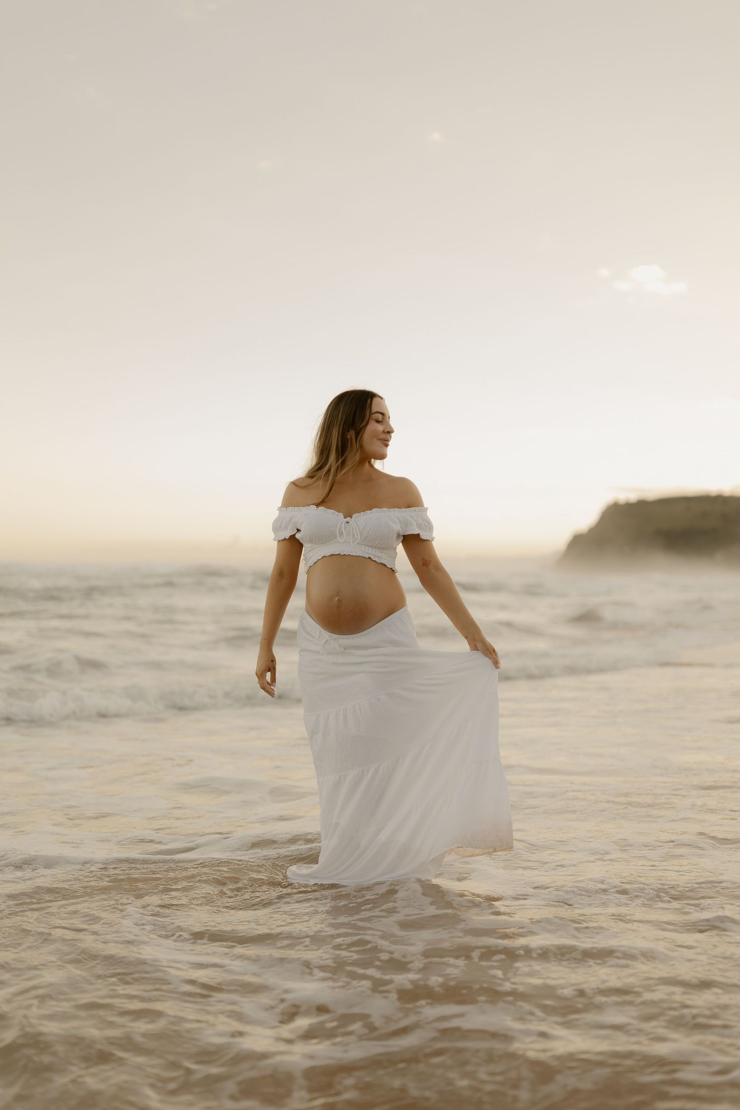 Woman in flowing white dress dancing on beach at sunset with waves in background.