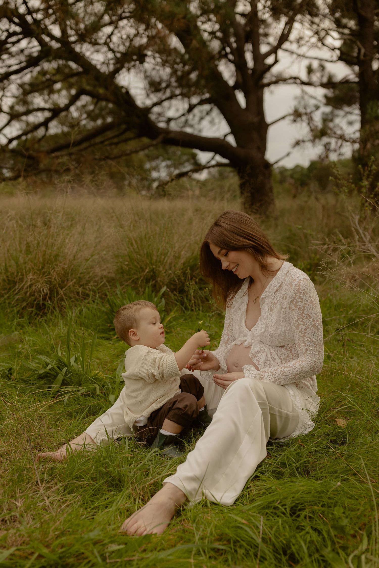 Pregnant mother in white lace dress sits on grass sharing a tender moment with her toddler outdoors.