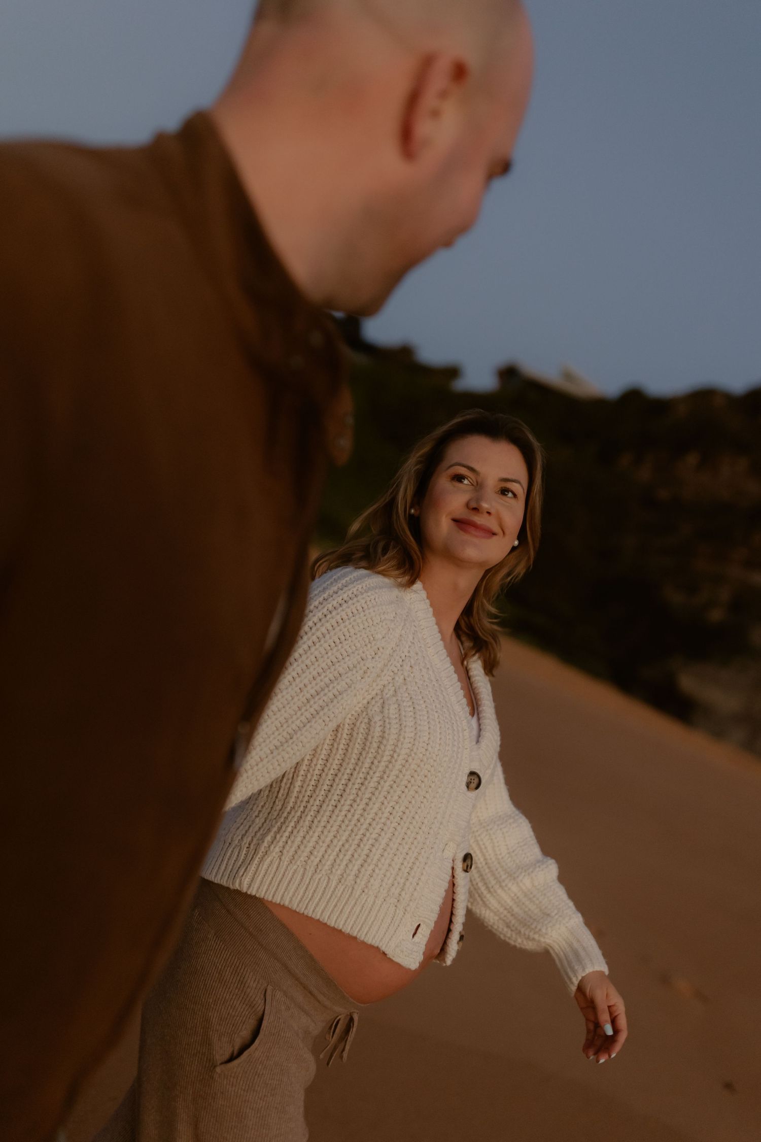 Pregnant woman in white knit cardigan smiling over shoulder on sandy beach at blue hour.