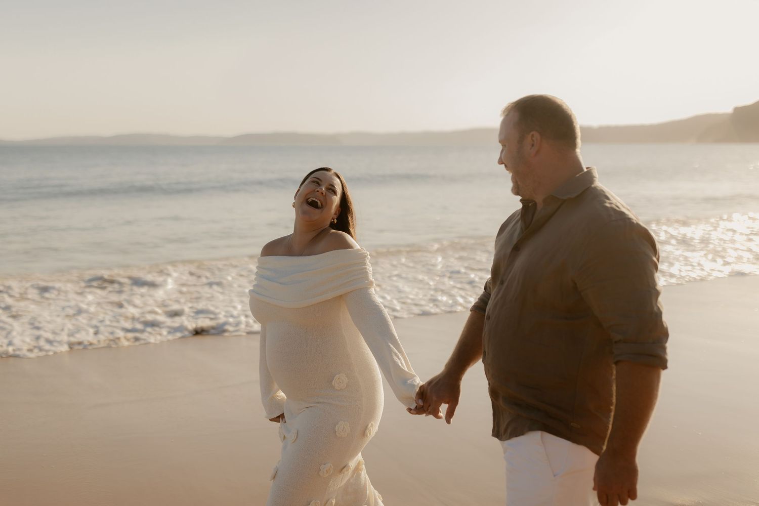 A couple walking and laughing together on a sunny beach at sunset.