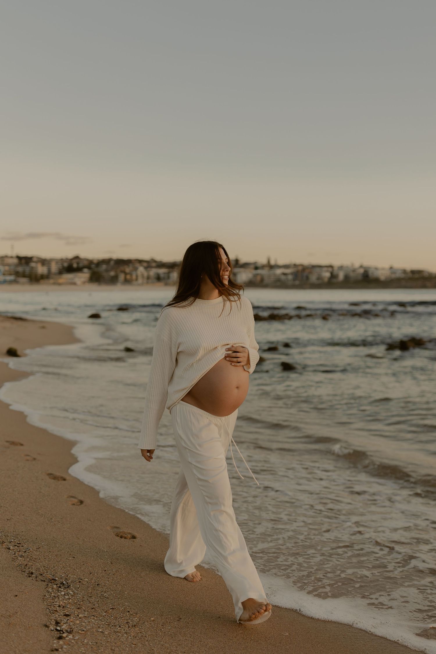 Expectant mother in flowing white outfit walks along beach at sunset with waves crashing on shore.