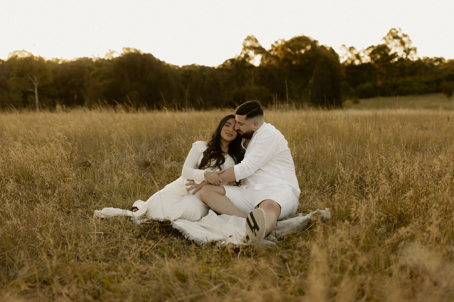 Intimate couple lounges together on blanket in rustic field during golden hour.