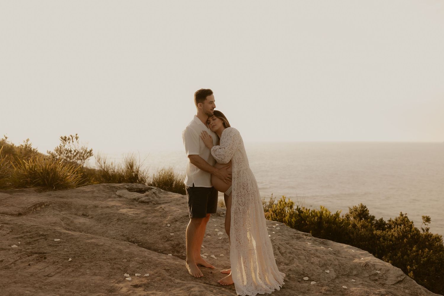 A couple in light colored clothing stands on a hilltop overlooking a scenic valley at sunset.