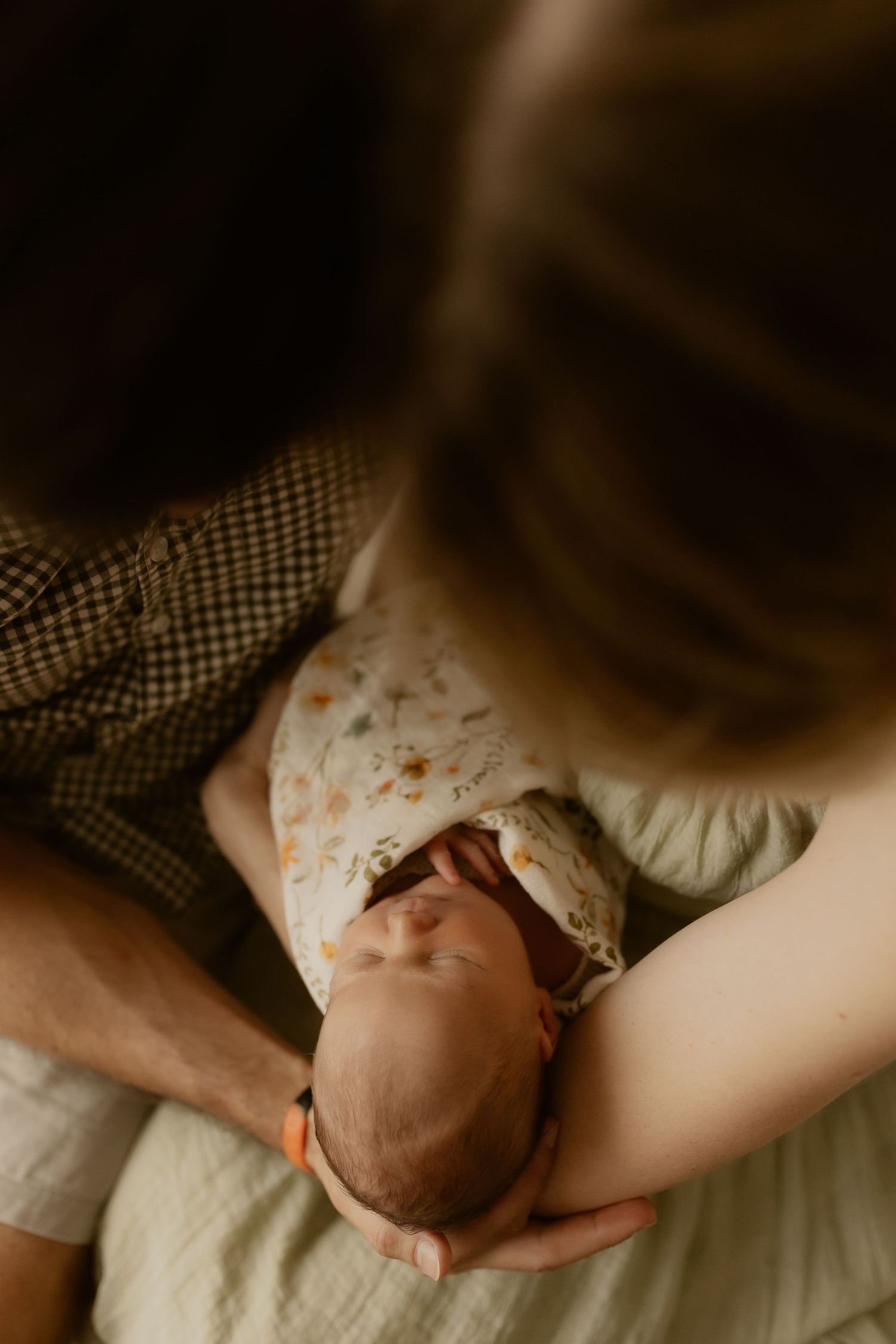 Gentle hands cradling a sleeping newborn in floral clothing against neutral background.