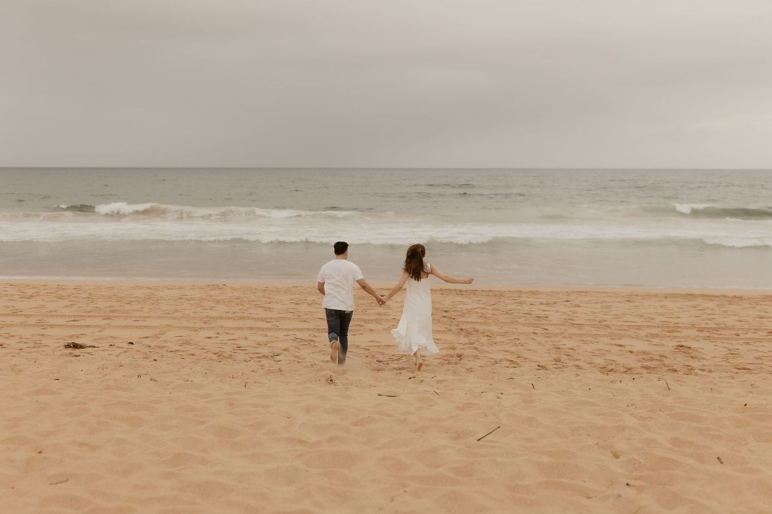 A couple walks hand in hand along a serene beach with soft muted tones.