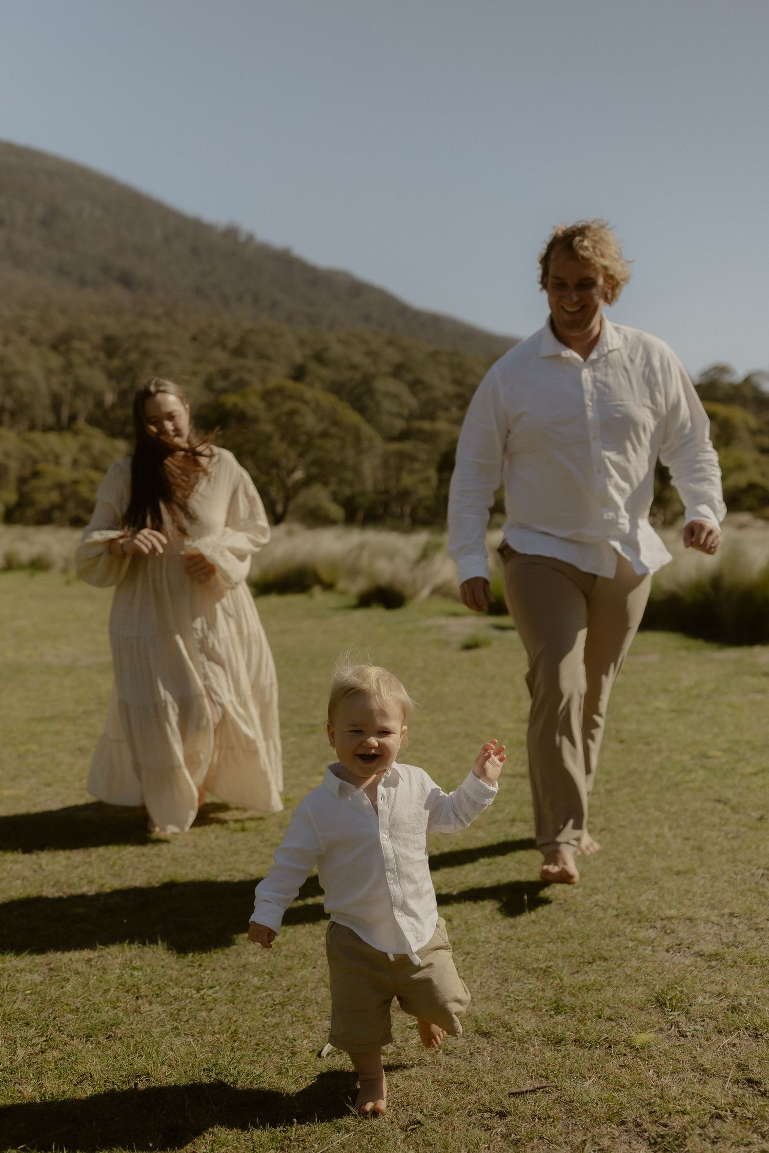 A family in white clothing runs and plays together in a grassy field on a sunny day.