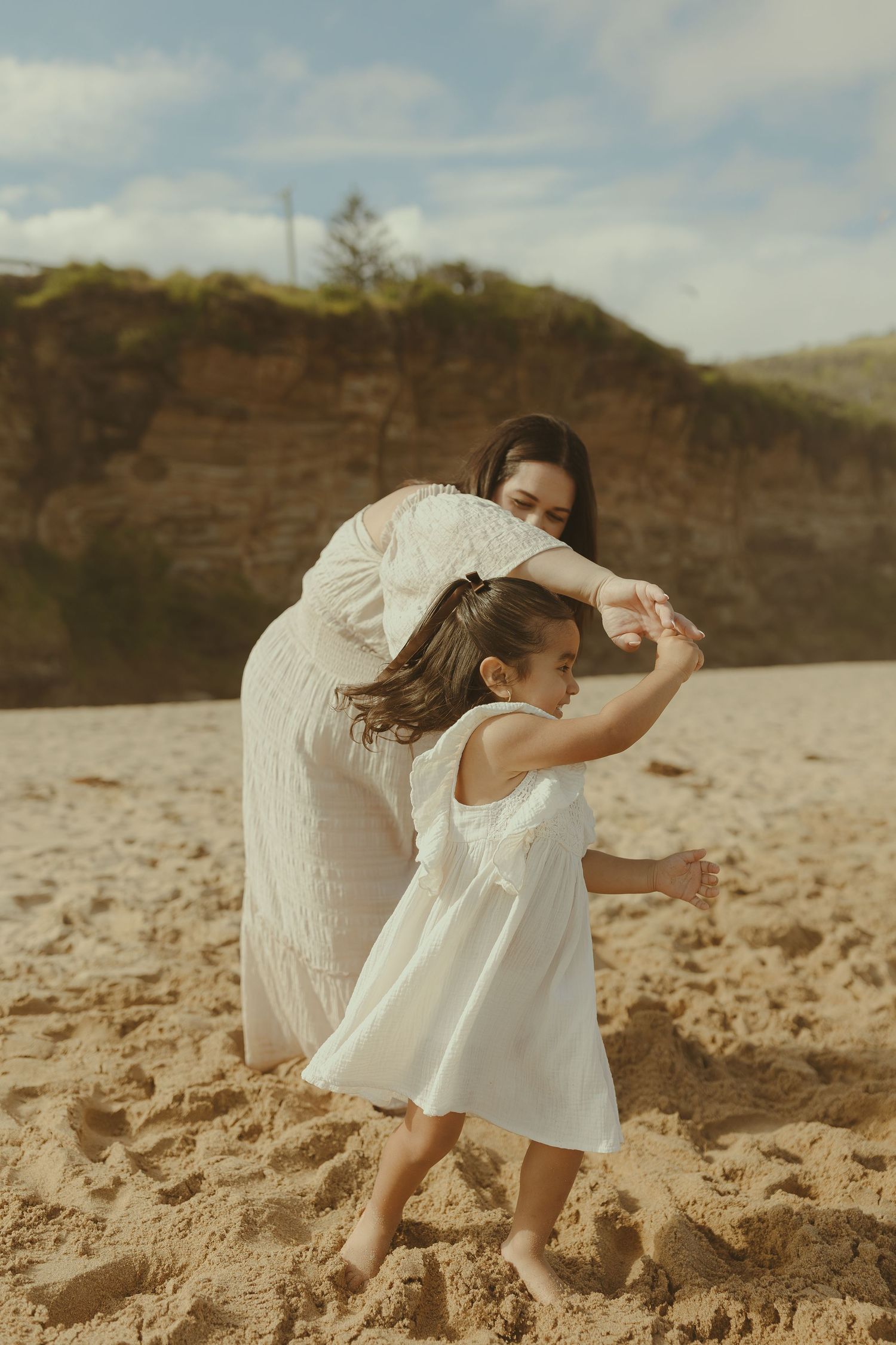 Two figures in flowing white dresses dance together on a sandy beach against a backdrop of rocky cliffs.