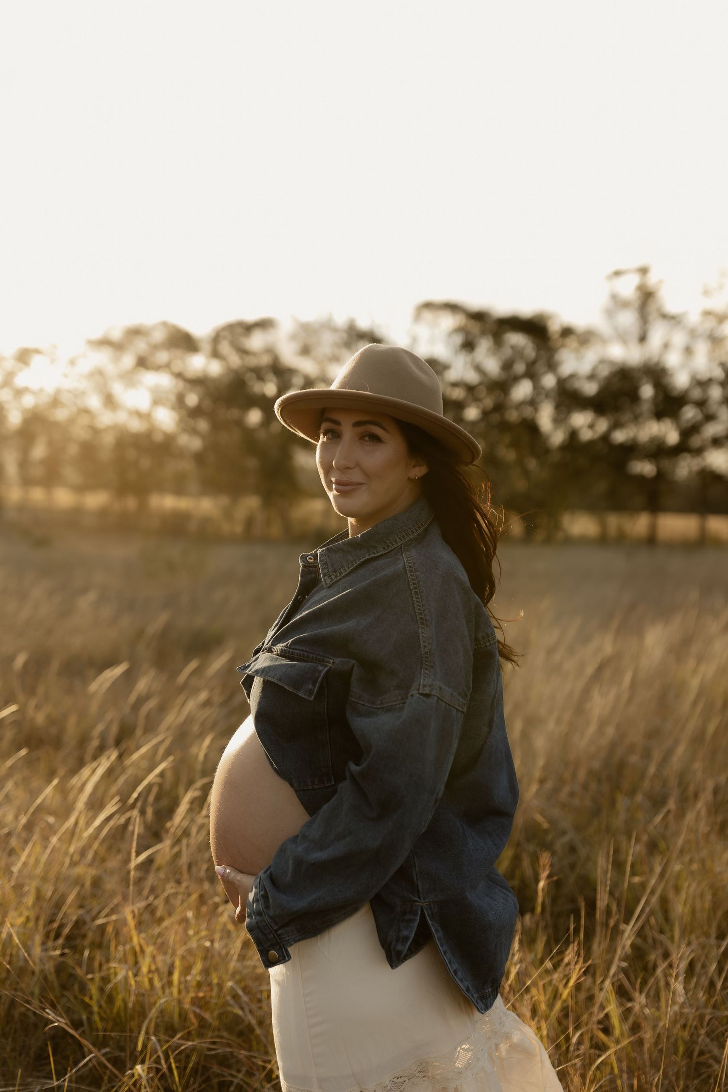 A maternity photo in a field at sunset shows an expectant mother in a denim jacket and wide-brimmed hat.