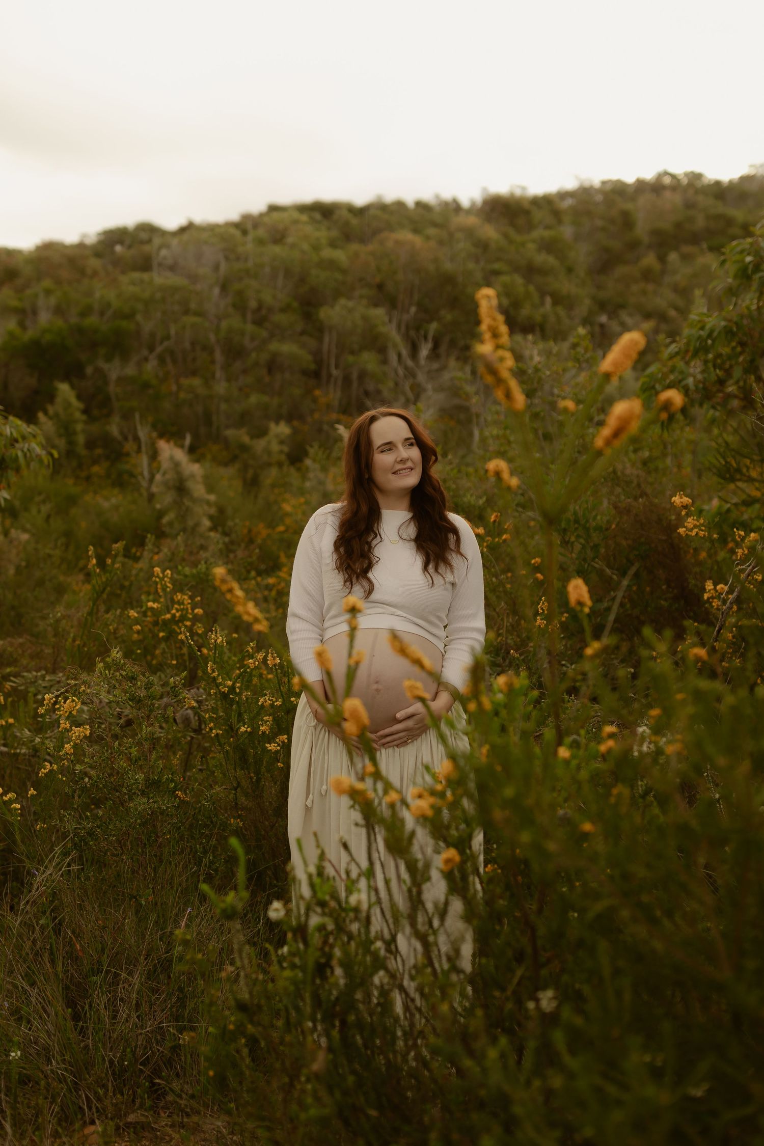 Pregnant woman in white dress standing in lush green field with yellow wildflowers during golden hour maternity photoshoot.