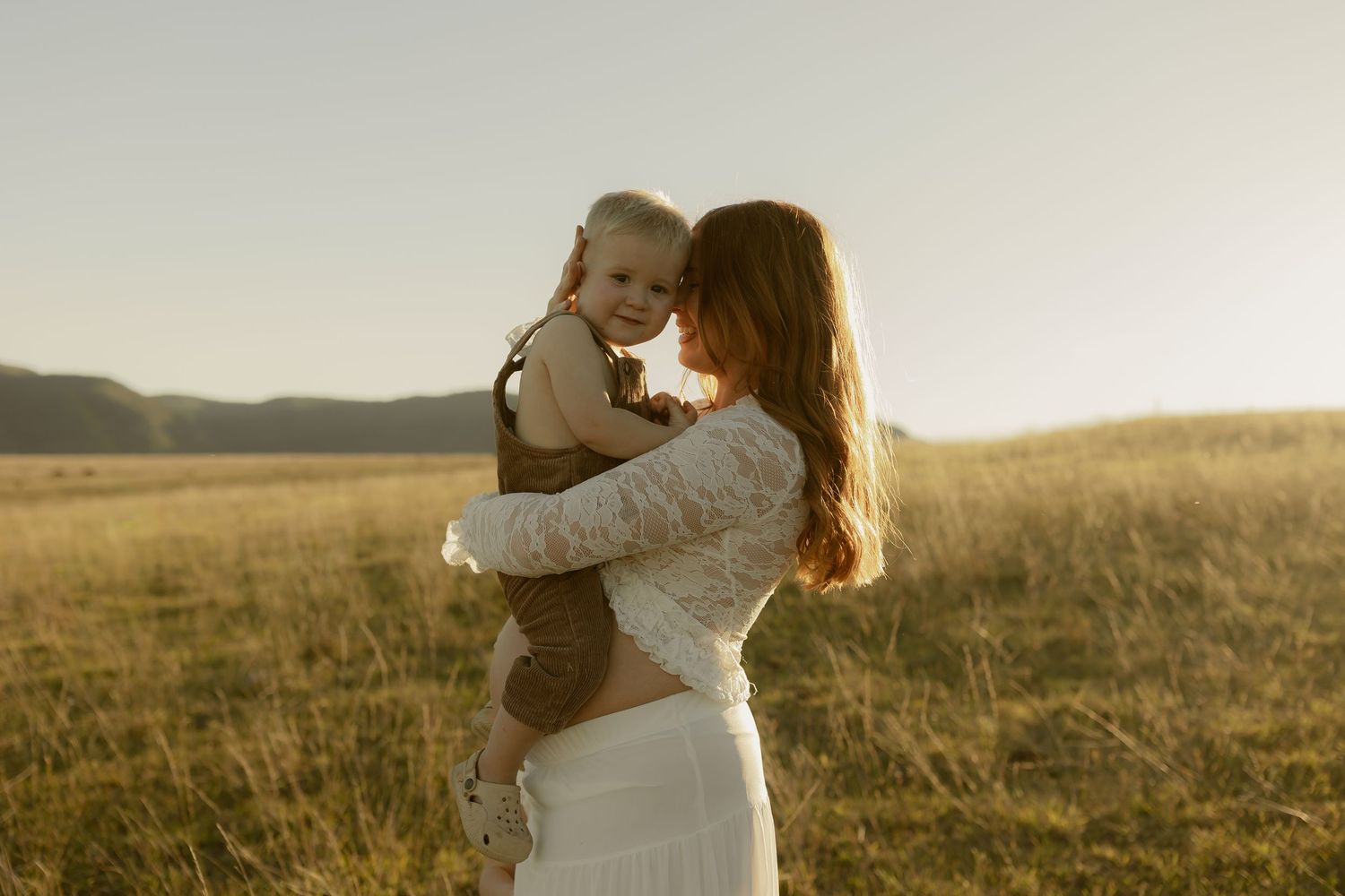 A mother holds her smiling baby in a golden sunlit field, wearing a white lace dress during a warm outdoor photo session.