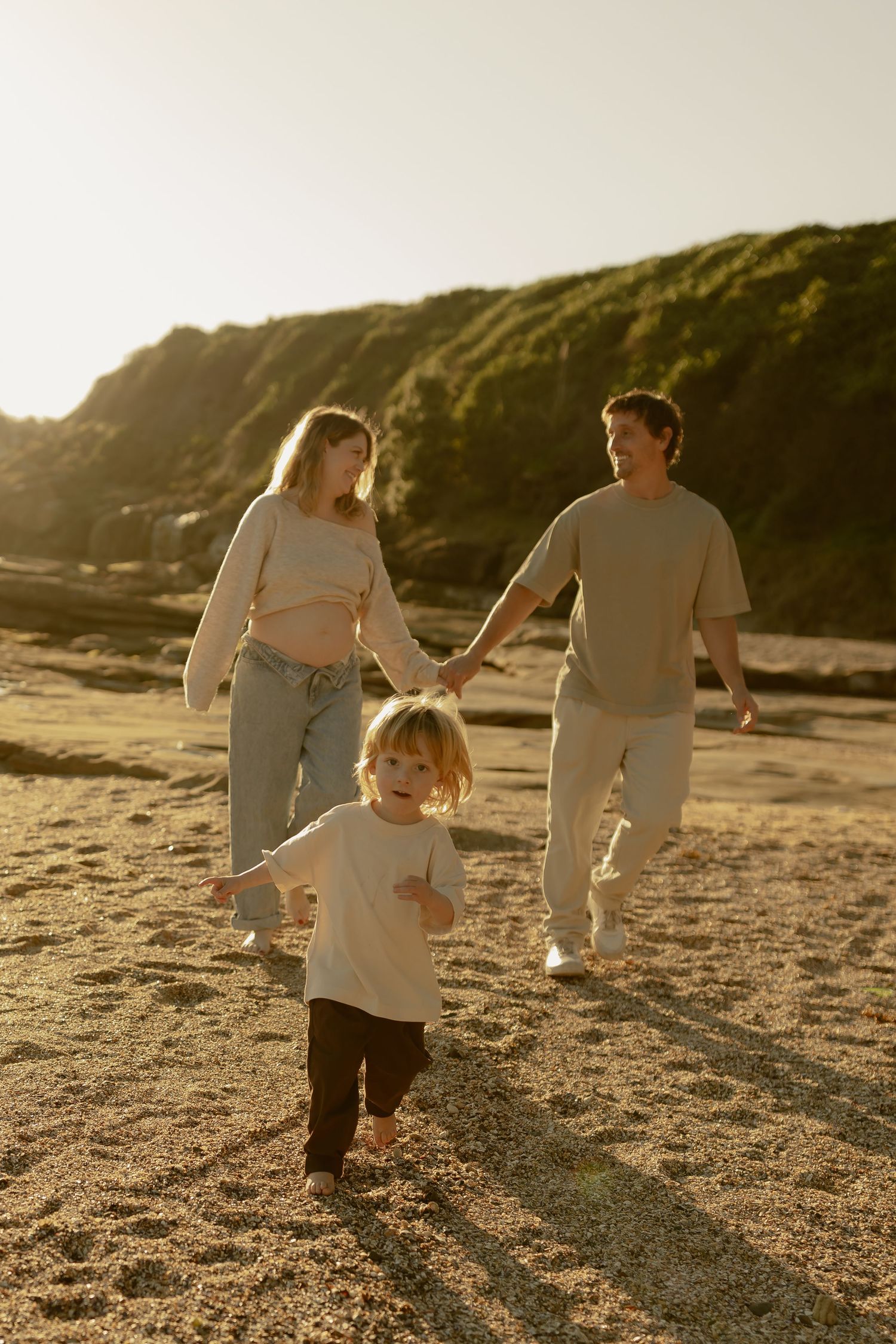Family walking on sandy beach at golden hour, parents holding hands with young child running ahead near rocky cliffs.