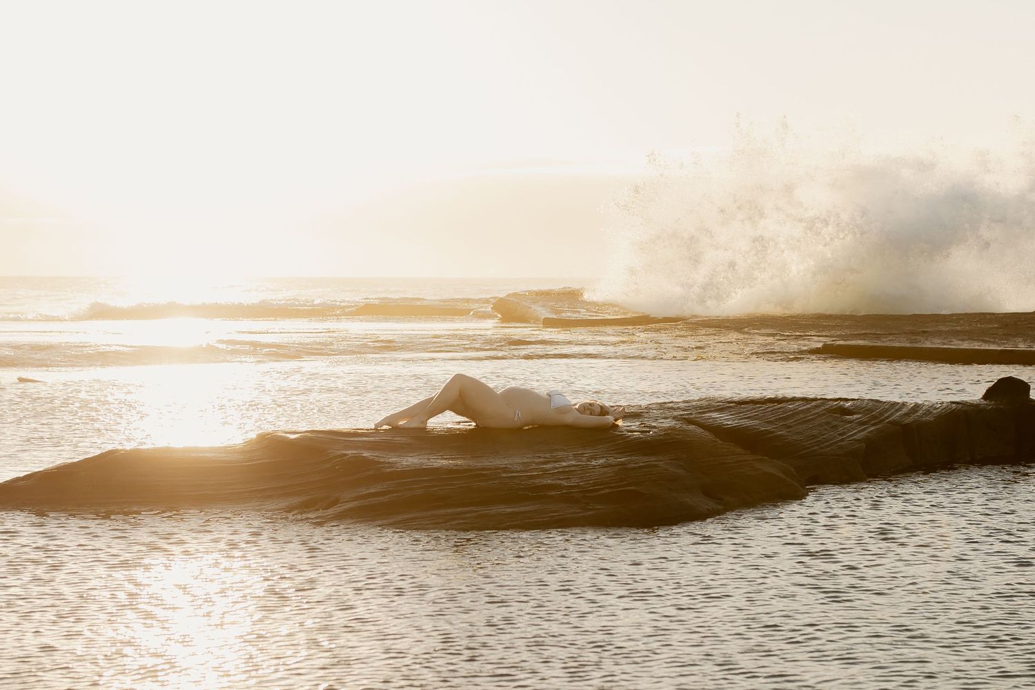 A panoramic seascape at sunset with warm golden light reflecting off calm ocean waters and distant mountain silhouettes.