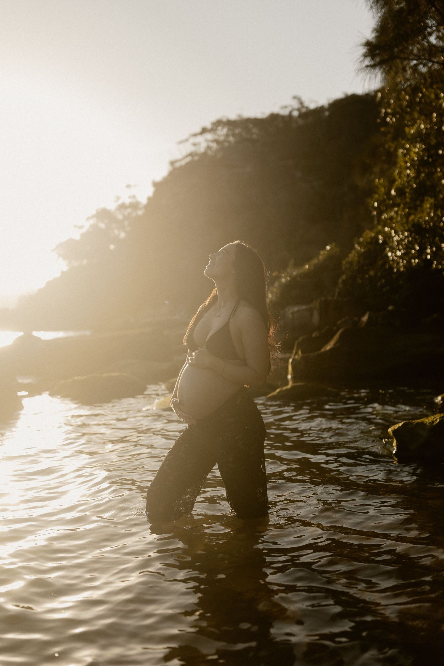 Silhouette of an expecting mother standing in water at sunset, creating a dreamy backlit maternity scene.