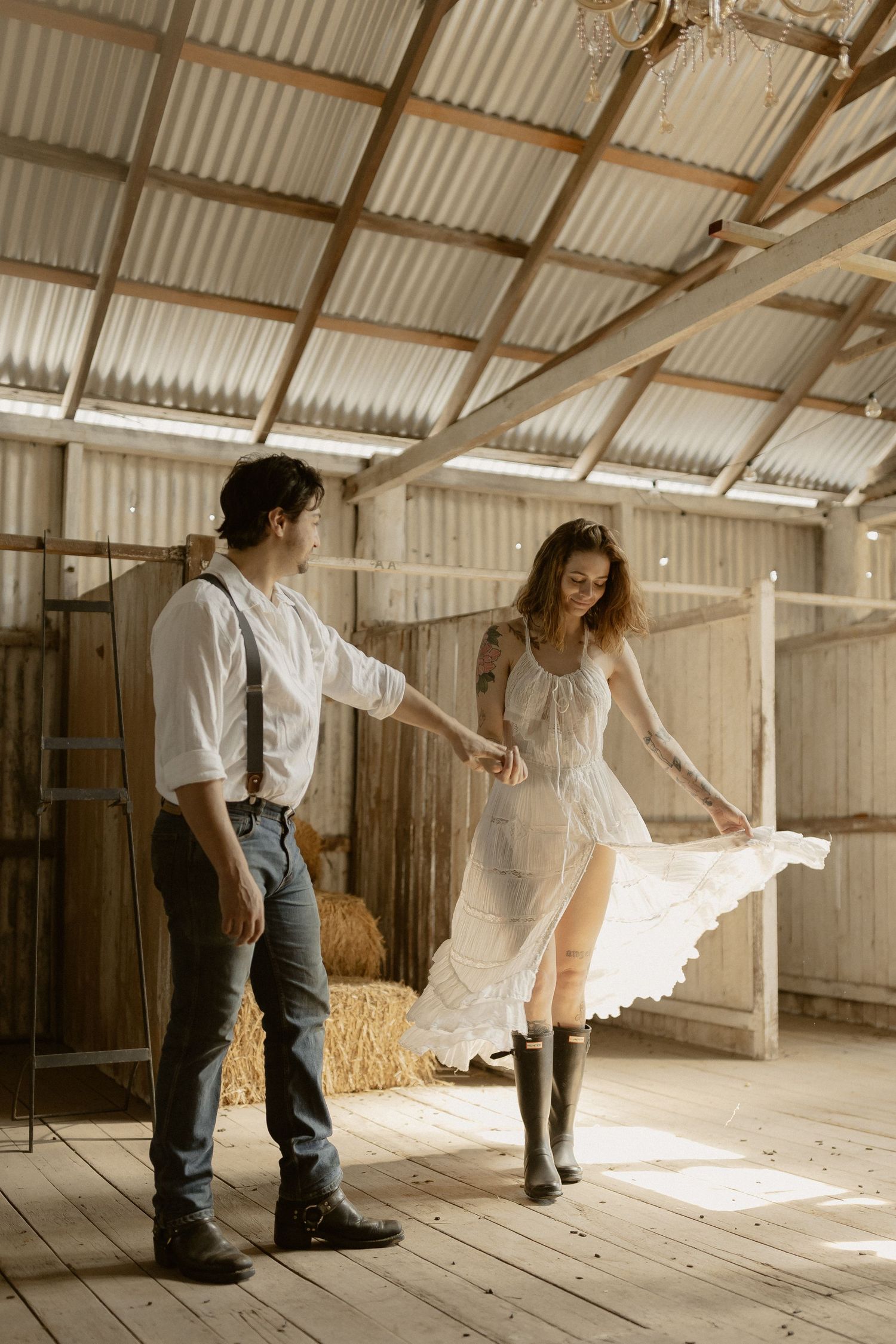 People dancing in flowing white clothes inside a rustic barn with corrugated metal roof and wooden floors.