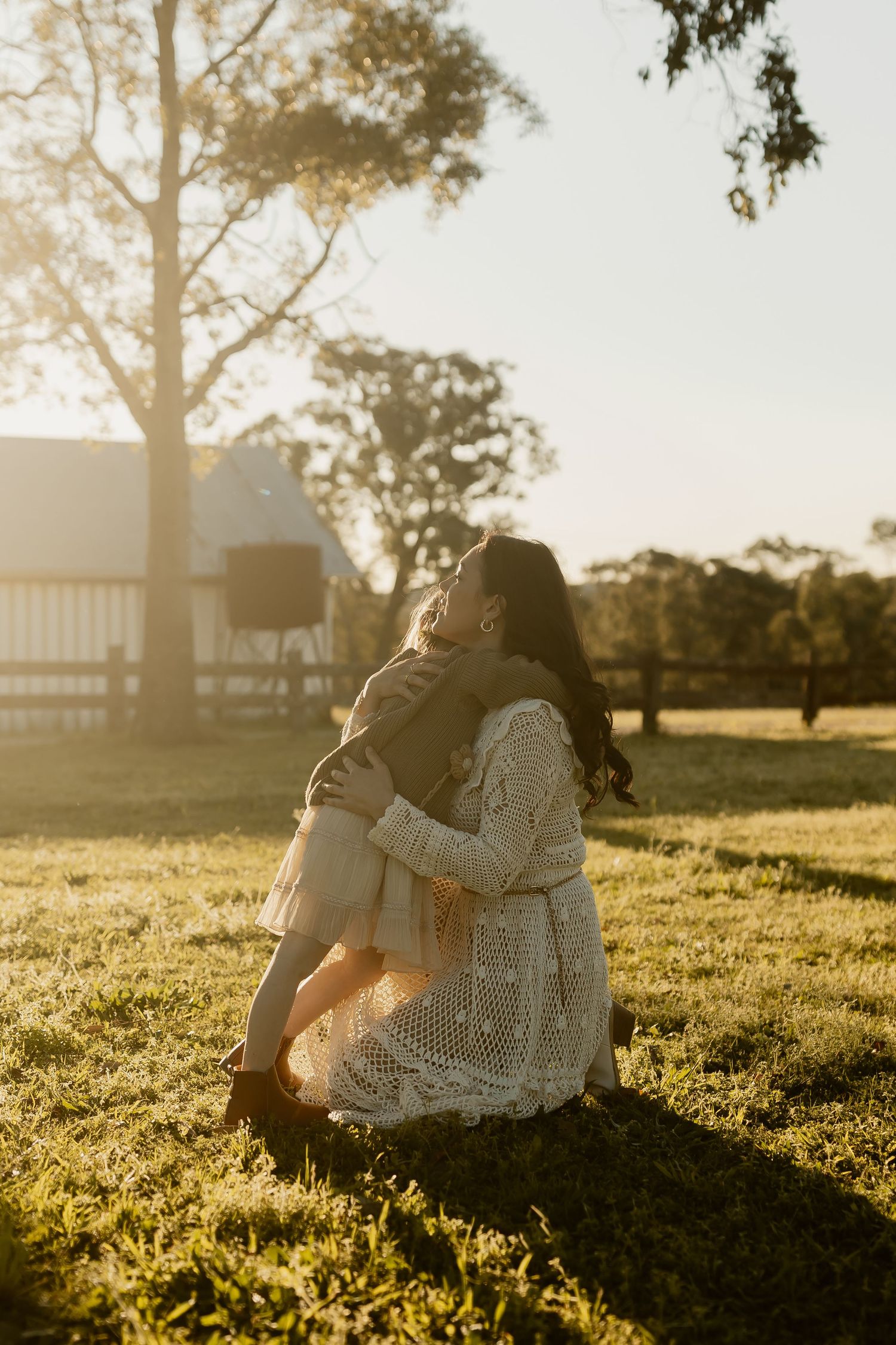A couple embraces on a grassy hillside during golden hour with trees silhouetted against the warm sunlight.