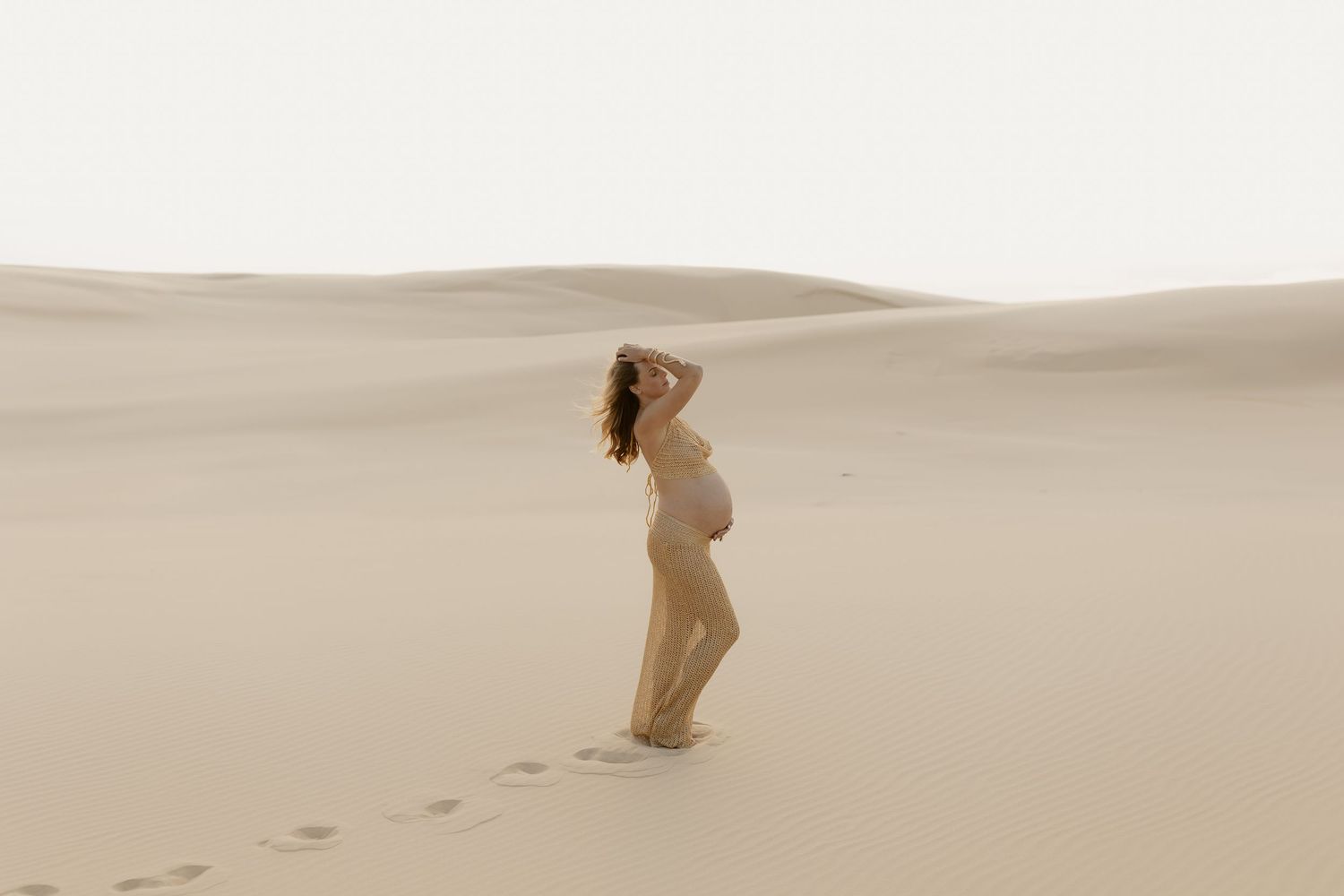 Person in light clothing walks across pristine sand dunes leaving footprints in the golden desert landscape.