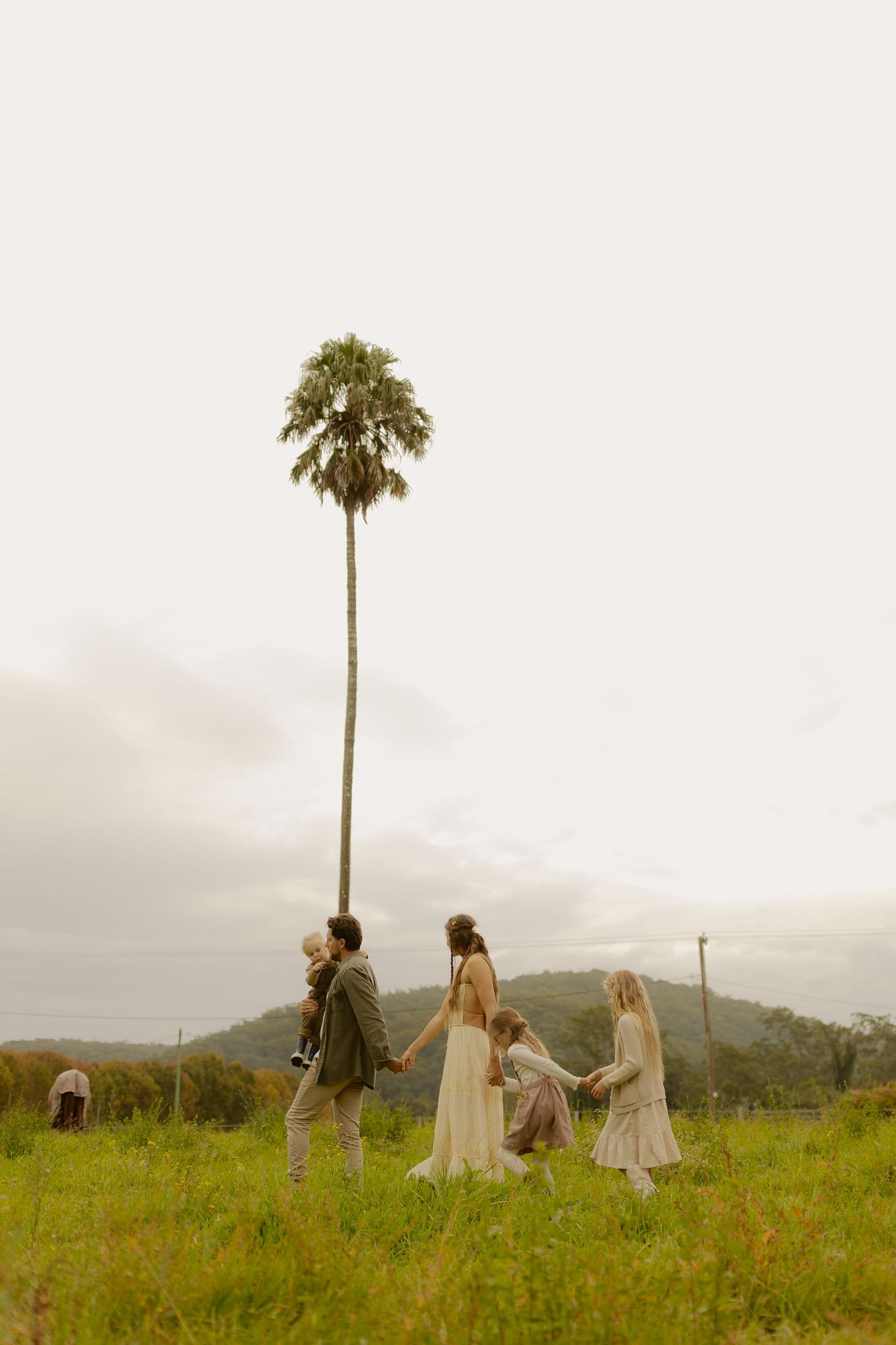 A family of five holds hands in a green field beneath a tall slender palm tree on a cloudy day.