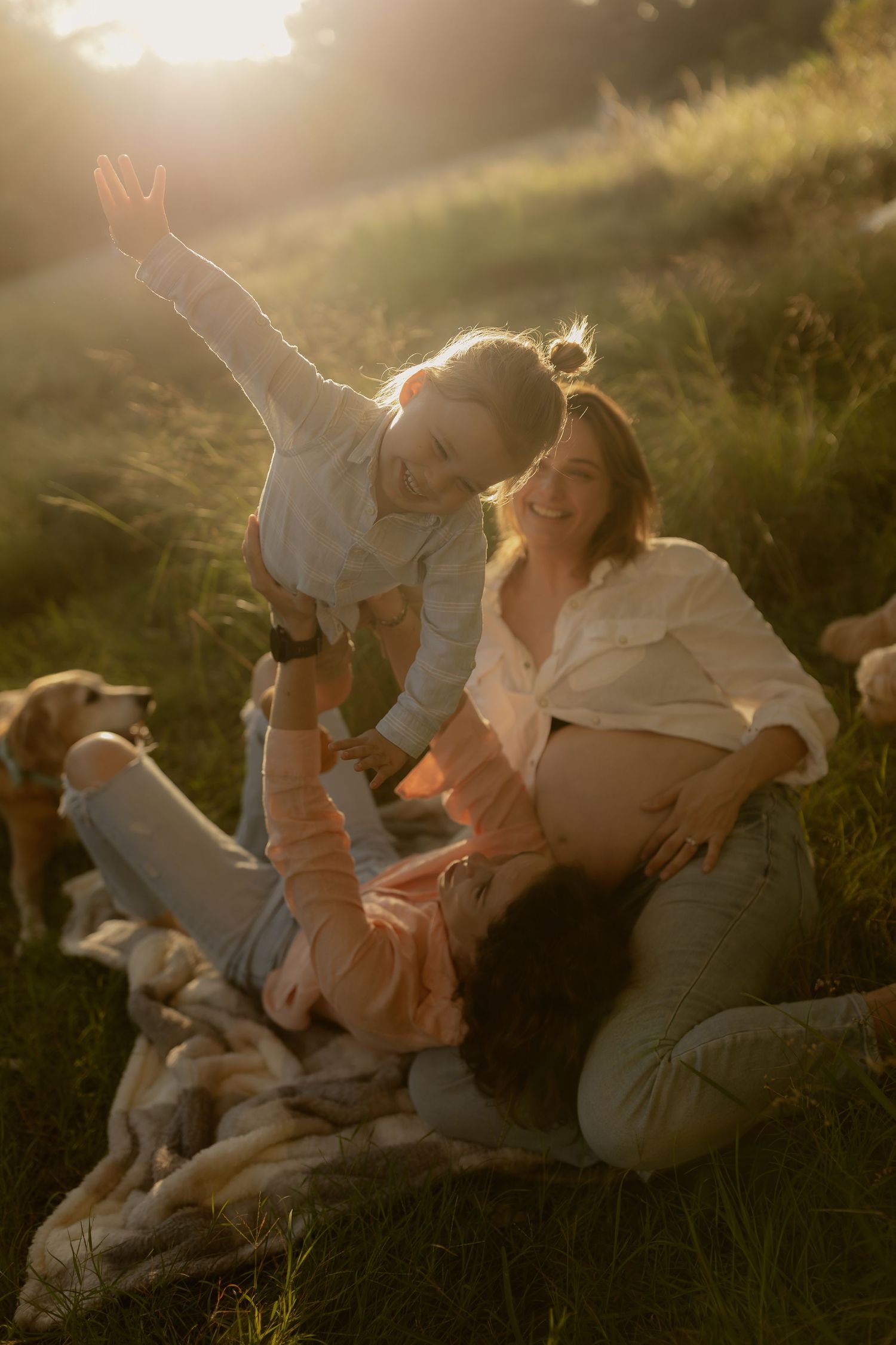Three women laughing and playing together on grass in golden sunset light with dogs resting nearby.