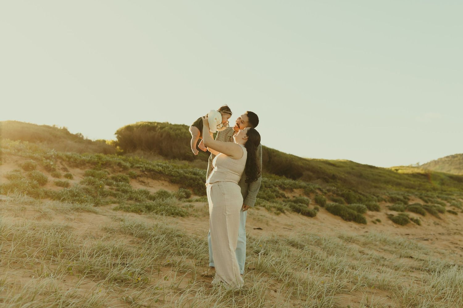 A couple in elegant white attire embraces on a windswept hillside during golden hour.