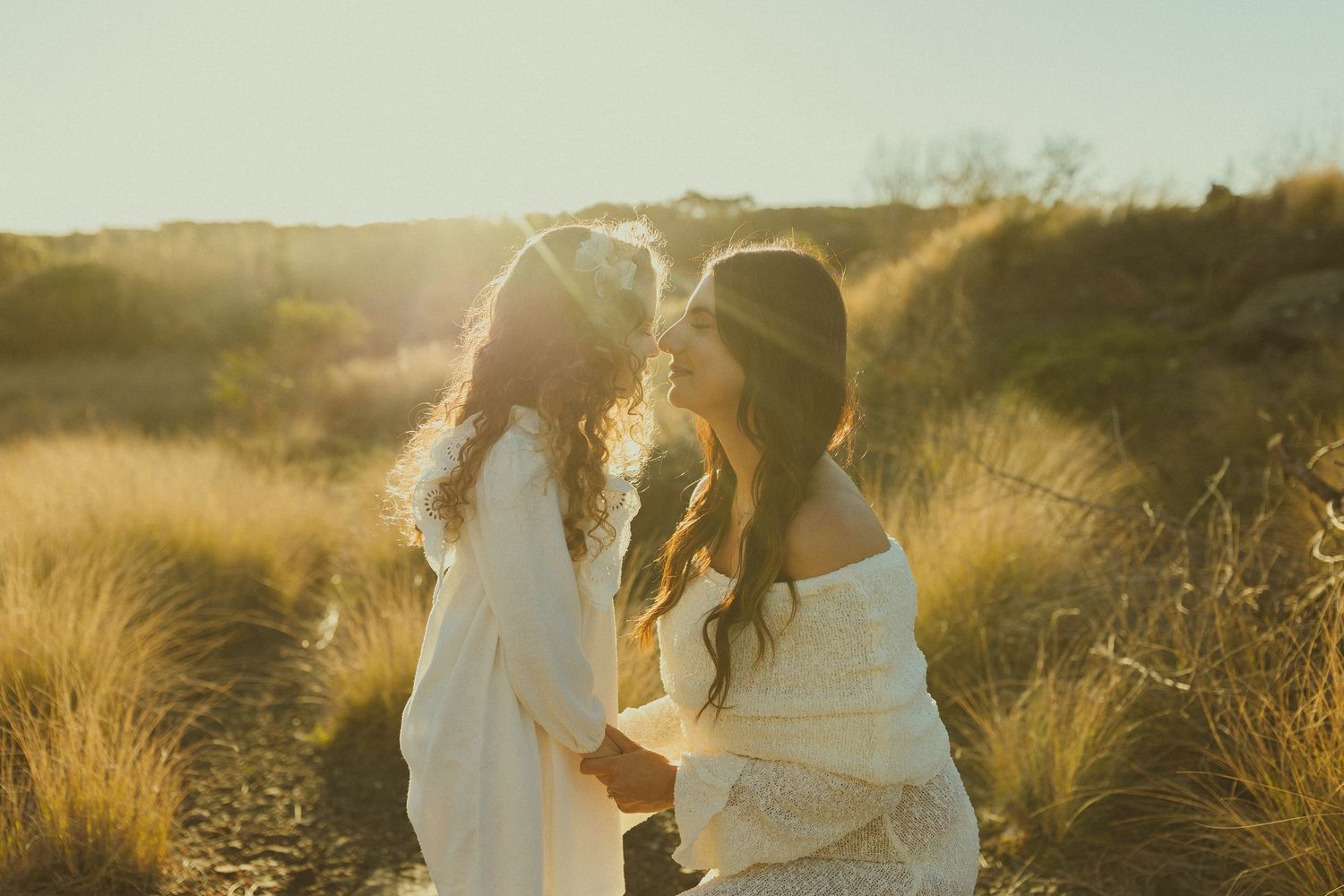 Two figures in white dresses standing together in golden sunlit field of tall grass at sunset.