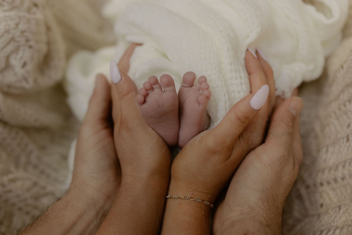 Two adult hands gently cradling tiny newborn baby feet wrapped in a soft cream knit blanket.