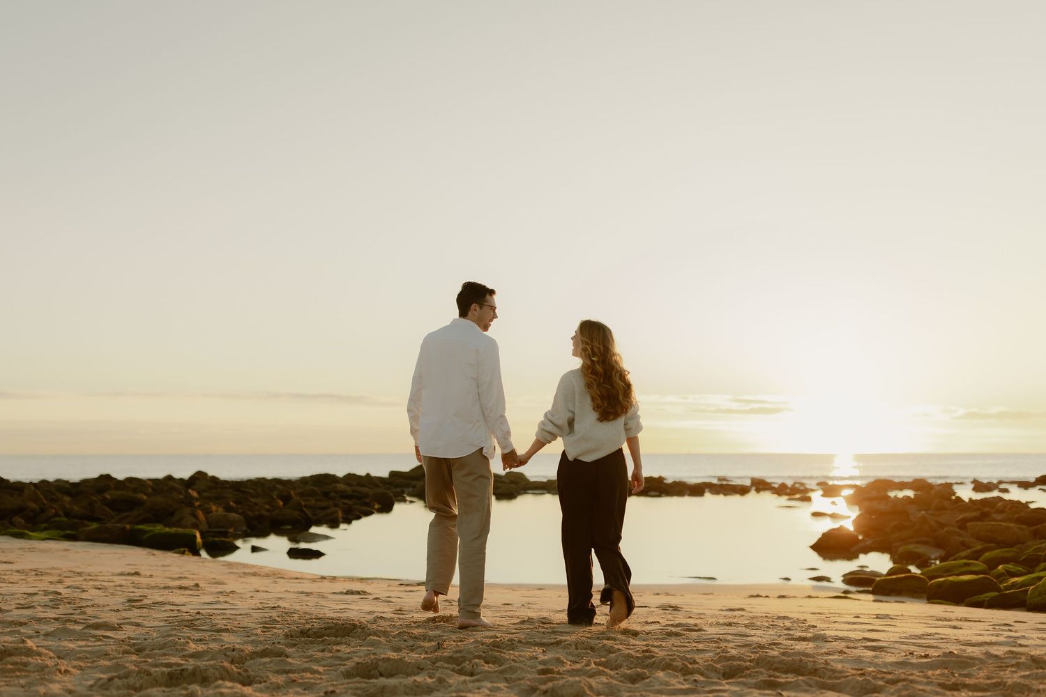 Couple holding hands on a sandy beach at sunset with rocky shoreline and golden sky backdrop.