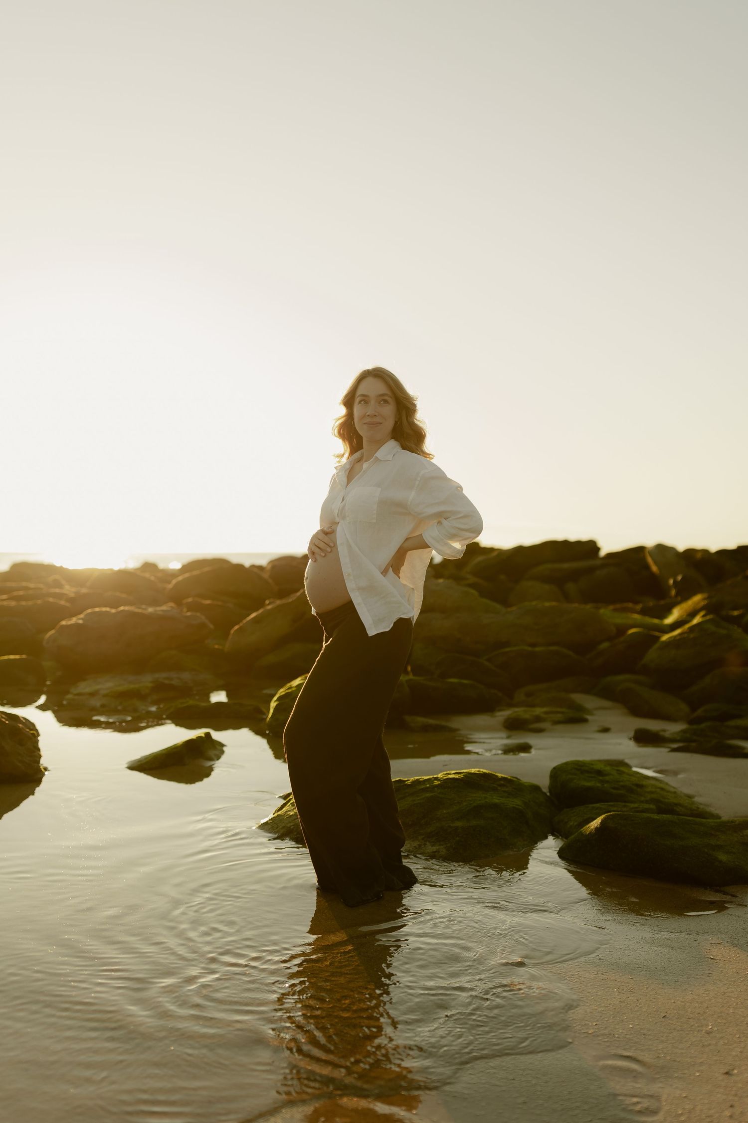 Pregnant woman in white shirt and black pants standing in shallow water among mossy rocks during golden hour sunset.